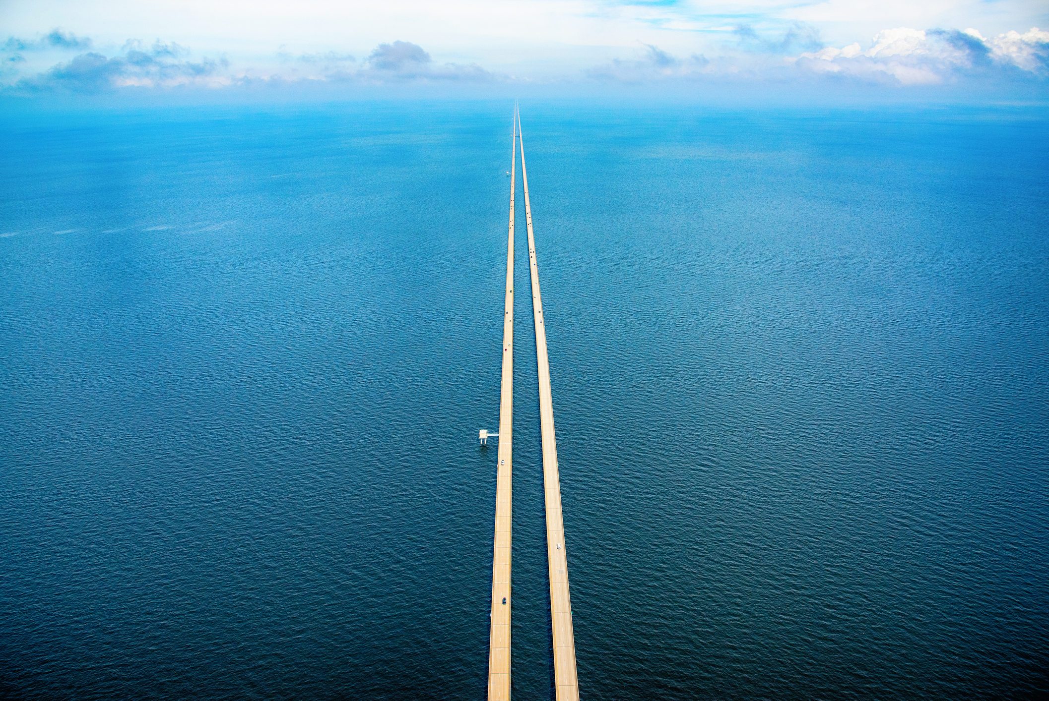 Long bridge stretches across calm blue water, viewed from above, with small vehicles traveling and clouds in the distant horizon.