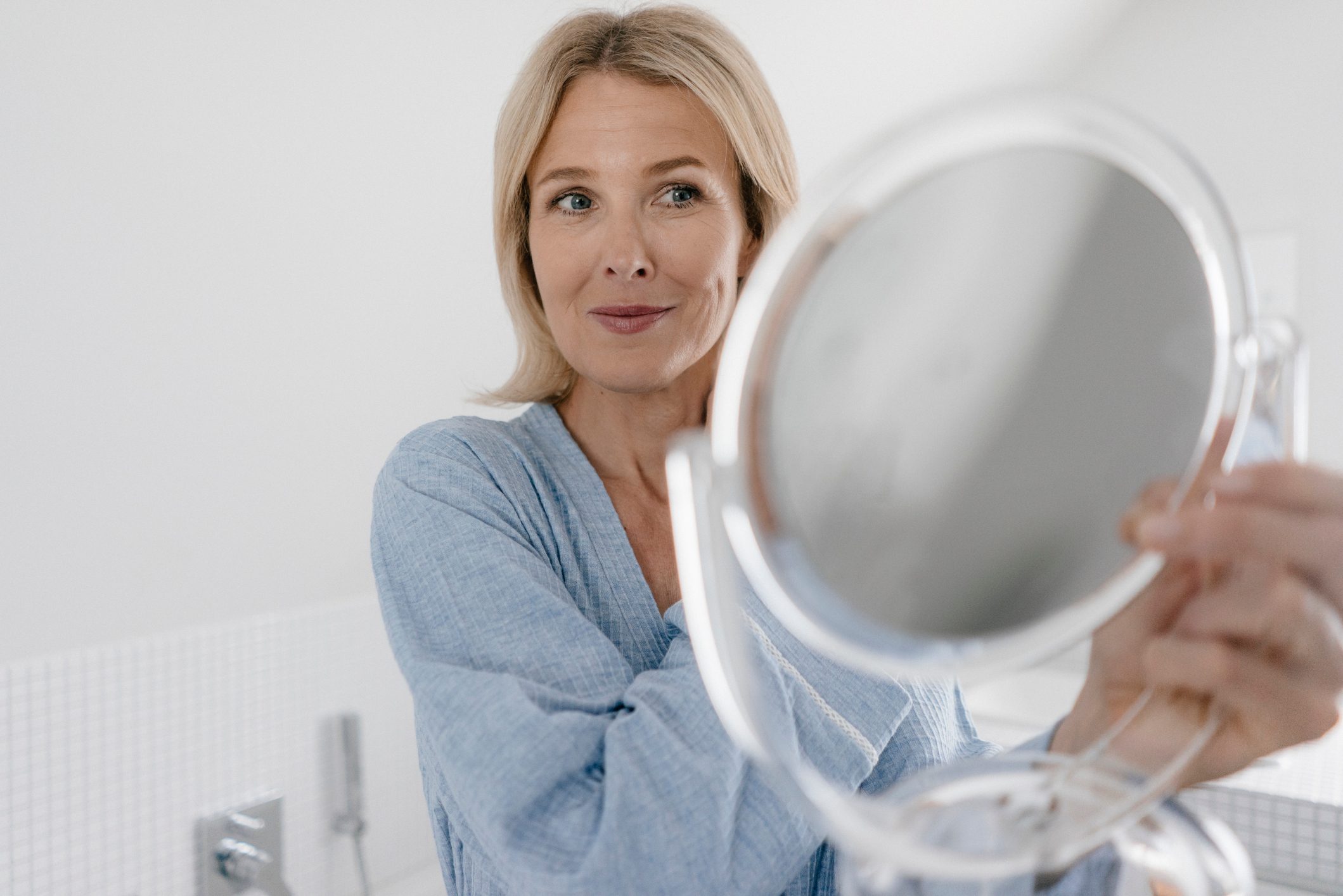 A person holds a round mirror, gazing into it, while standing in a bright bathroom wearing a light blue robe.