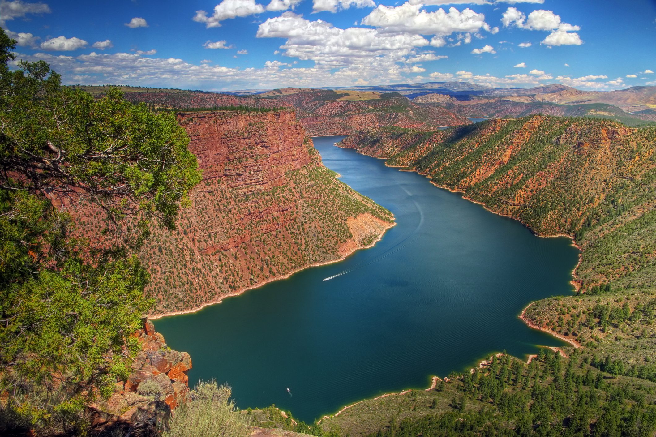 River winds through red canyon cliffs under a bright sky dotted with clouds, surrounded by dense greenery and rugged terrain.