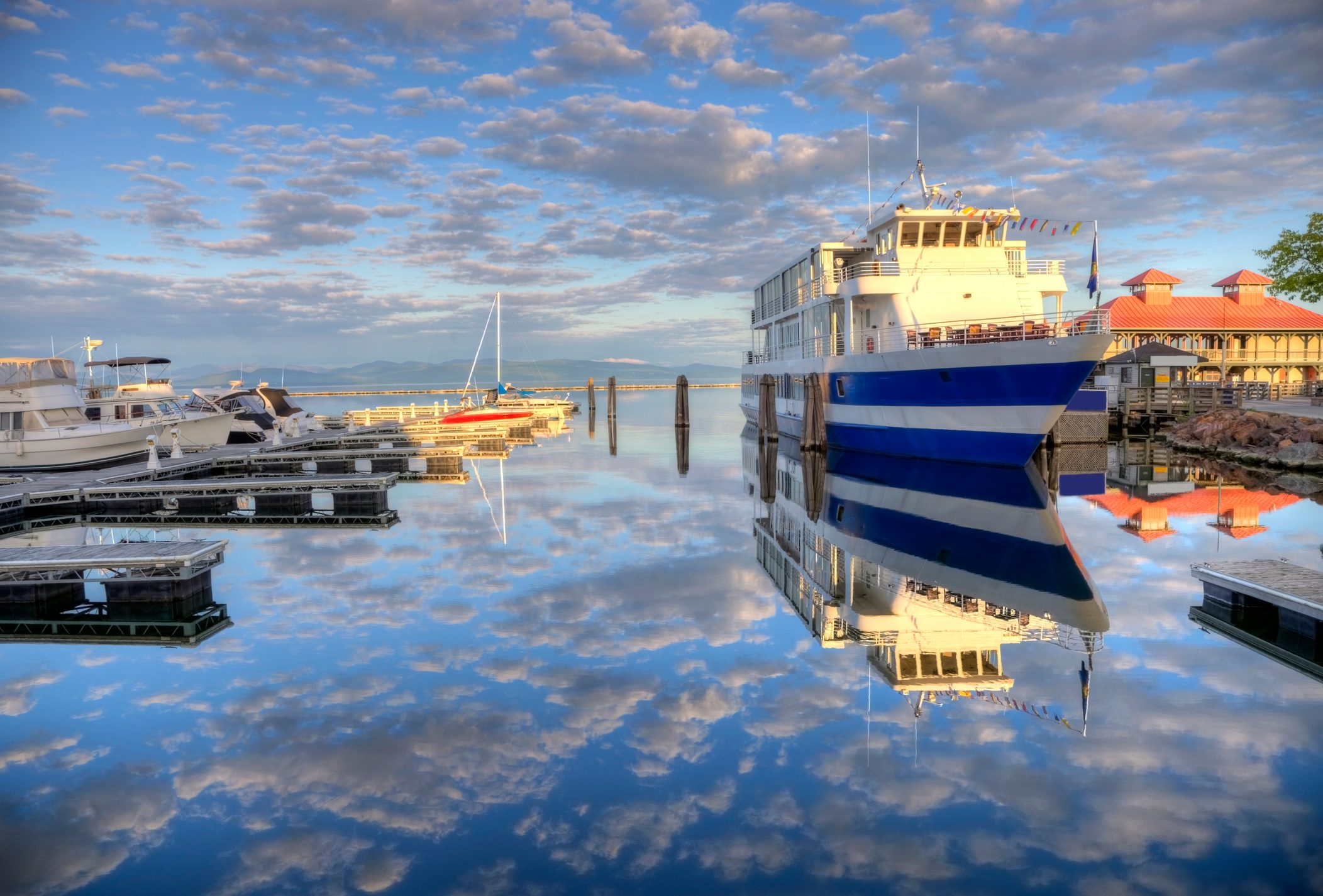 A large ferry sits docked, reflecting on calm water amidst a marina, under a partly cloudy, blue sky.