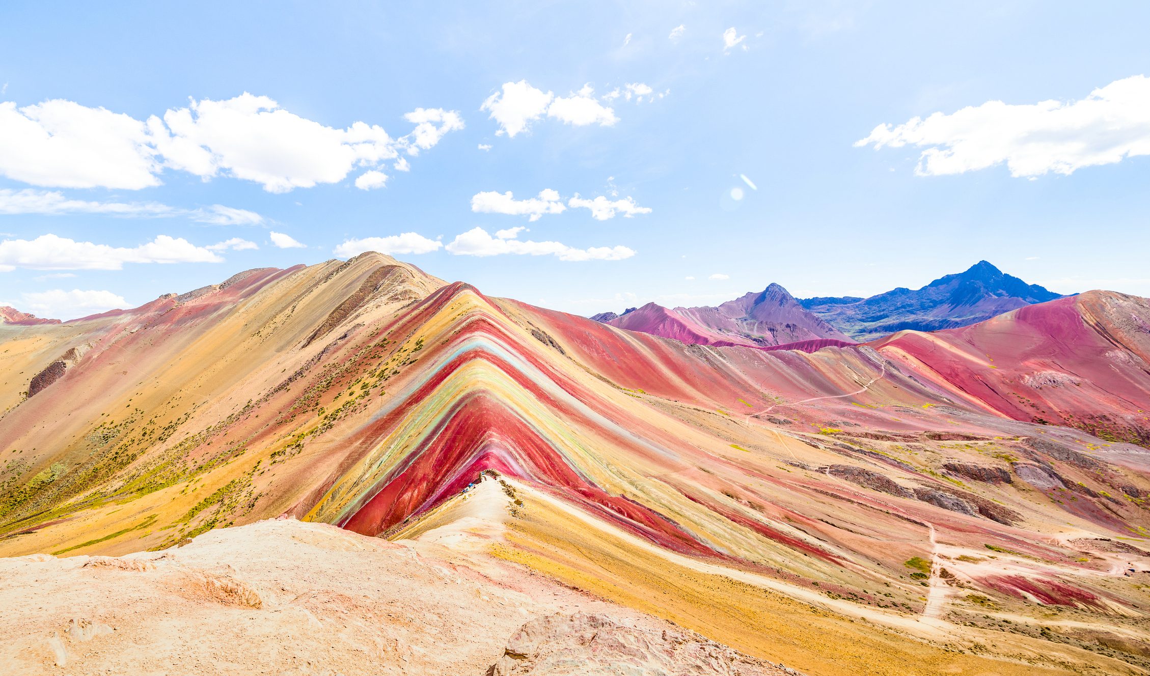 Colorful, layered mountain runs diagonally; vibrant hues of red, yellow, and green form stripes. Surrounding peaks rise under a clear blue sky with clouds.