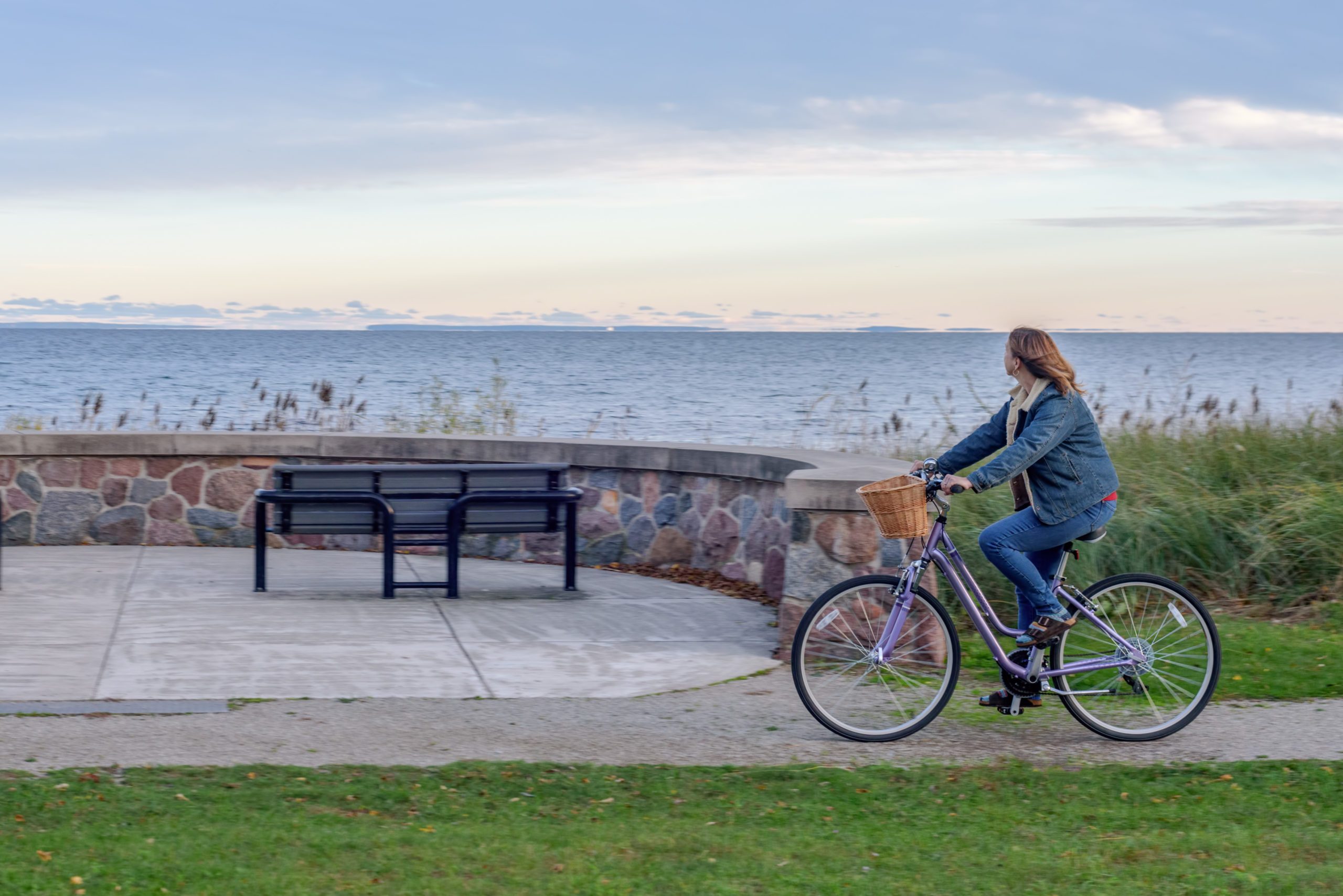 A person rides a bicycle along a coastal path near a stone wall and benches, with the ocean in the background.