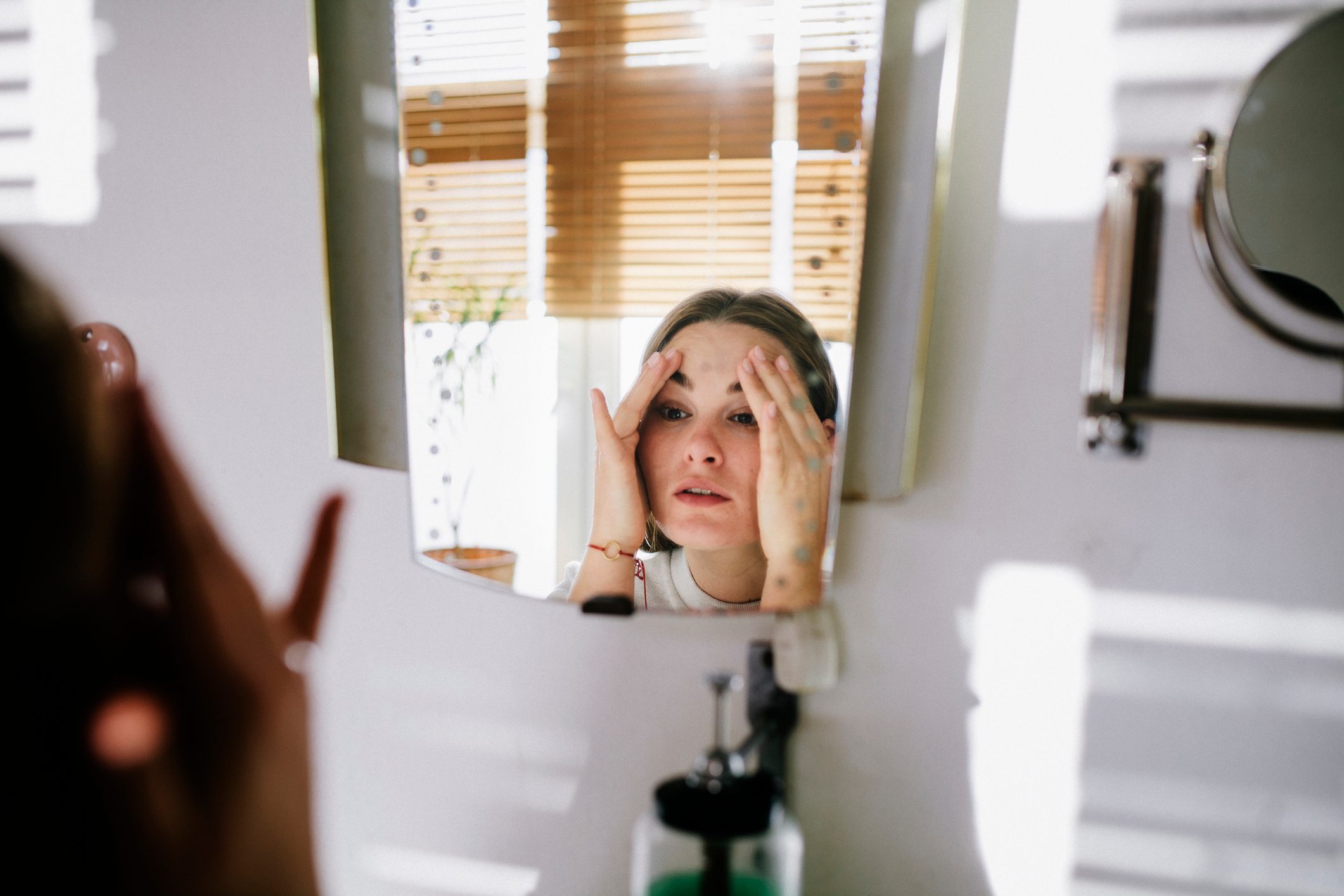 Person examining face in mirror; adjusting skin; room with sunlight filtering through blinds.