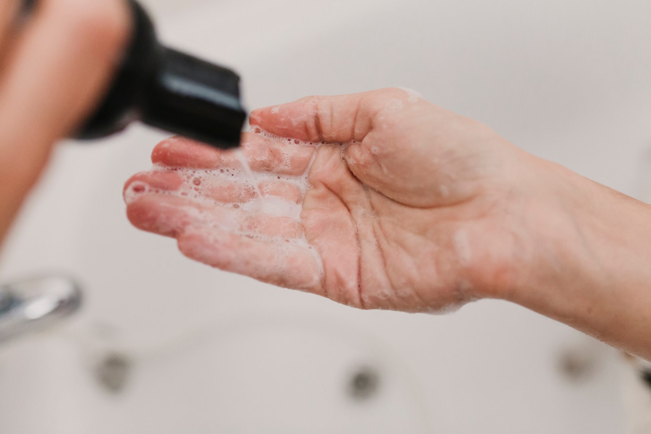 Hand receives foamy soap from a black dispenser, set against a blurred bathroom background.