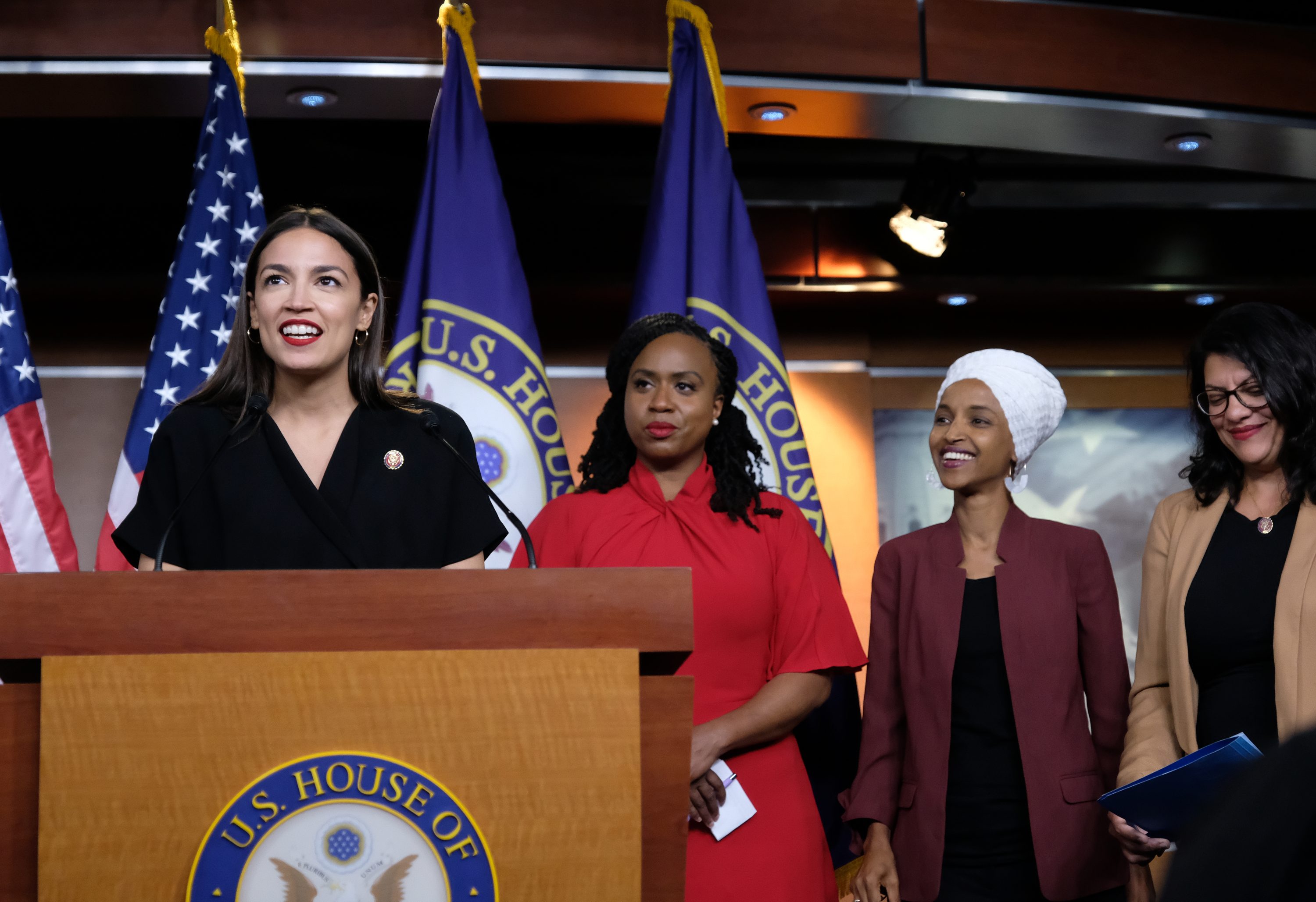 Four women stand, speaking and smiling, at a podium with "U.S. House of Representatives" insignia. American flags and House seals are in the background.