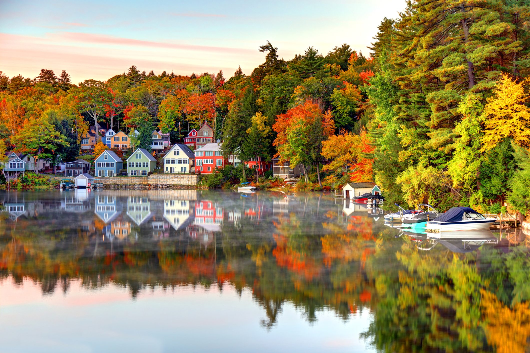 Colorful houses reflect on a calm lake amid autumn trees, with boats lined along the shore, creating a serene lakeside scene.