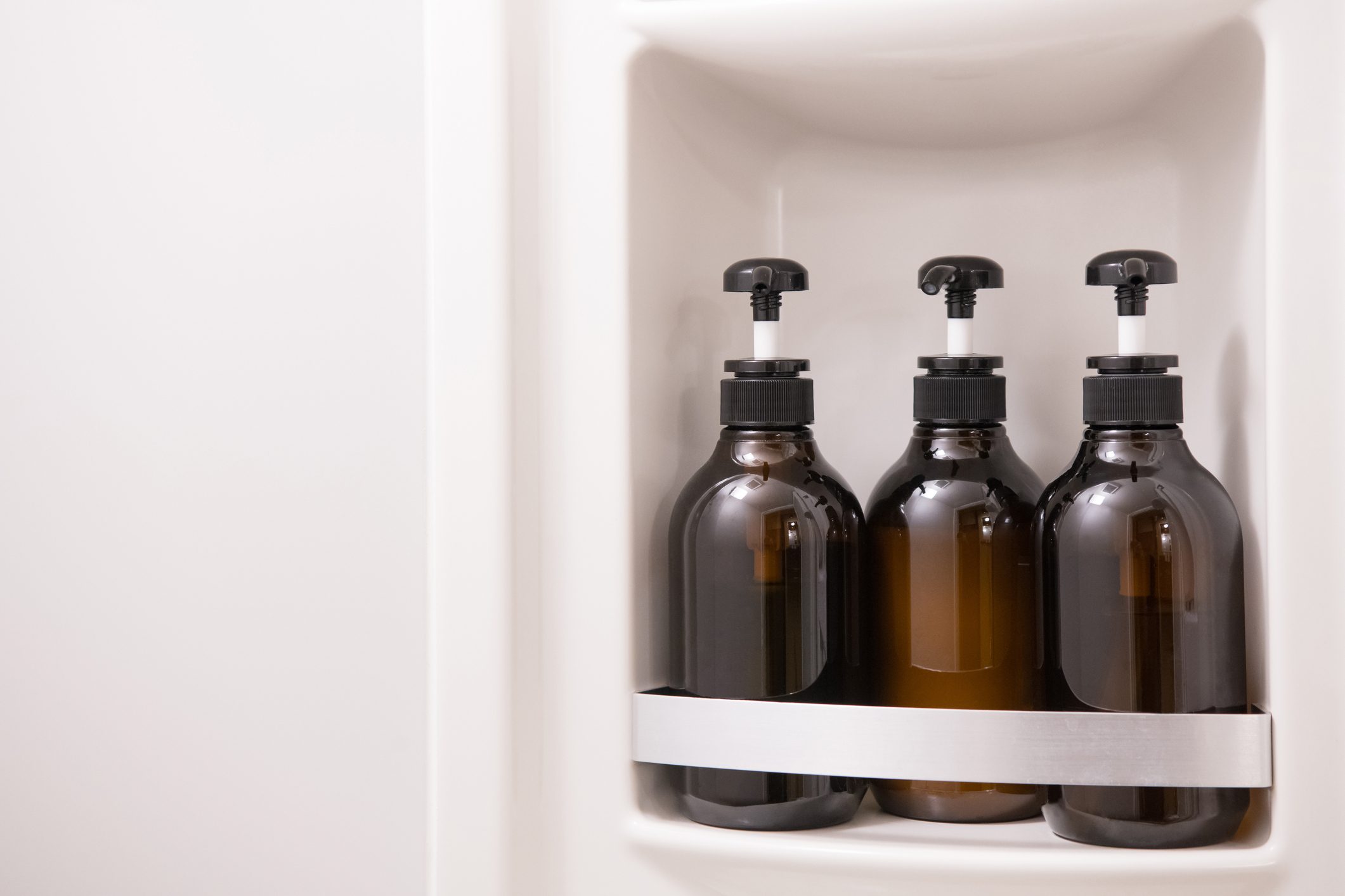 Three brown pump bottles sit in a white shower alcove, secured by a metallic holder.