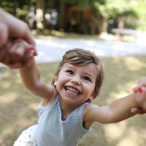 Child smiling, being swung by hands in a sunny park setting.
