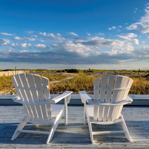 Two white chairs facing a grassy field, set on a wooden deck under a blue sky with scattered clouds.