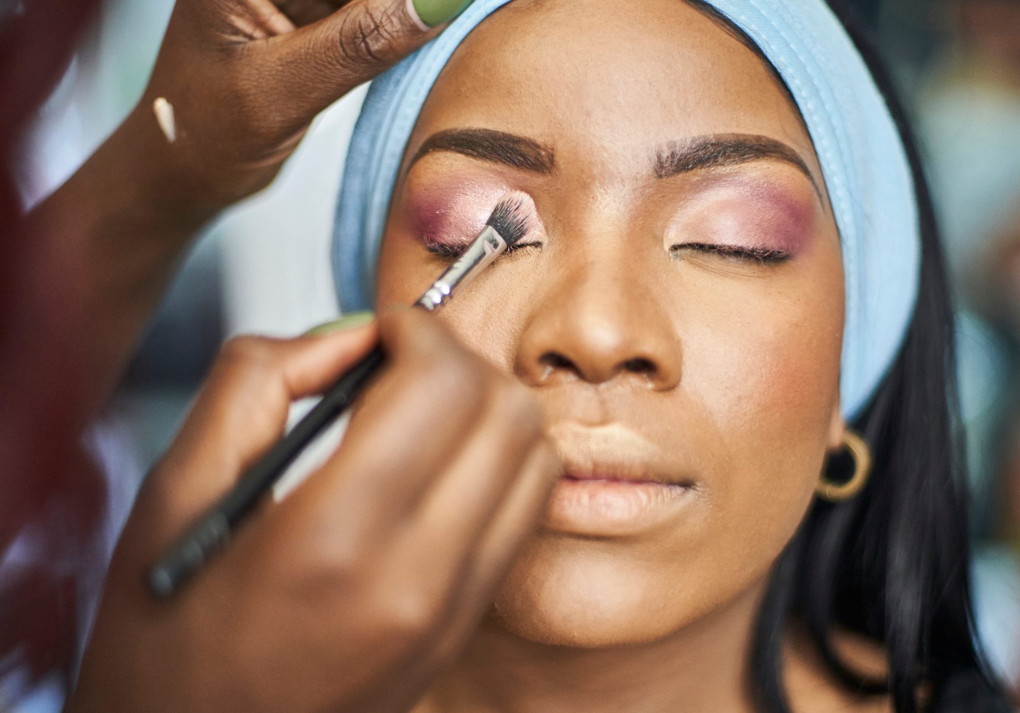 Face receiving makeup application, a brush applying eyeshadow on closed eyelid, framed by a blue headband in a close-up setting.