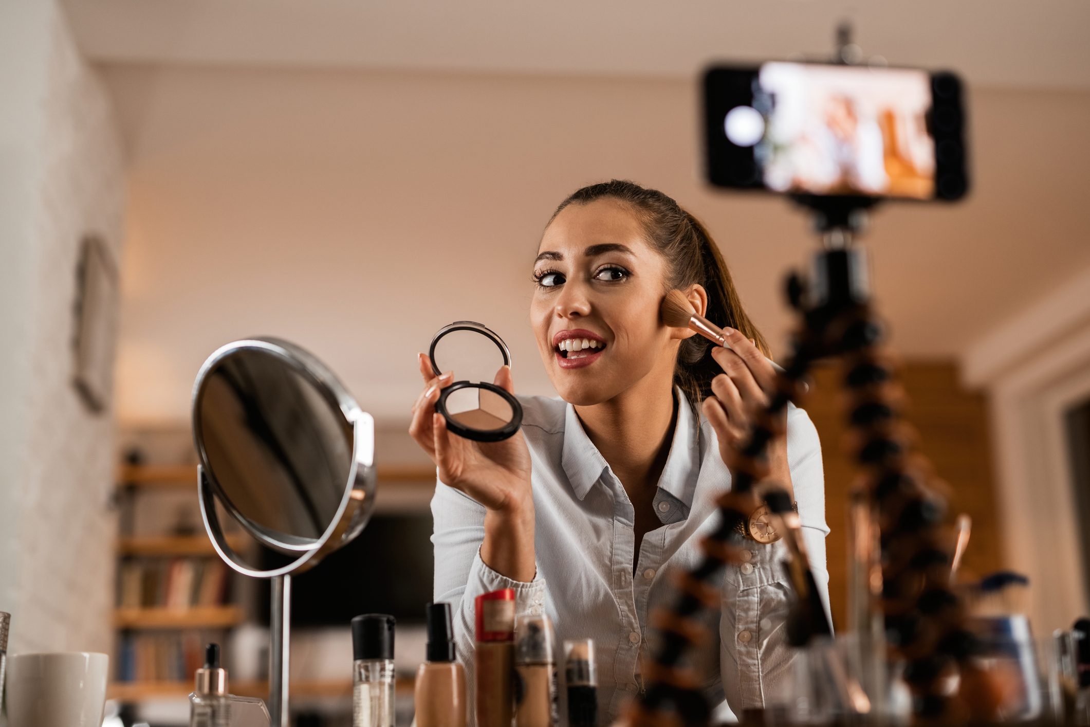 Person applying makeup, using a brush and compact, filming content. Set in a room with a mounted phone and various cosmetics on the table.