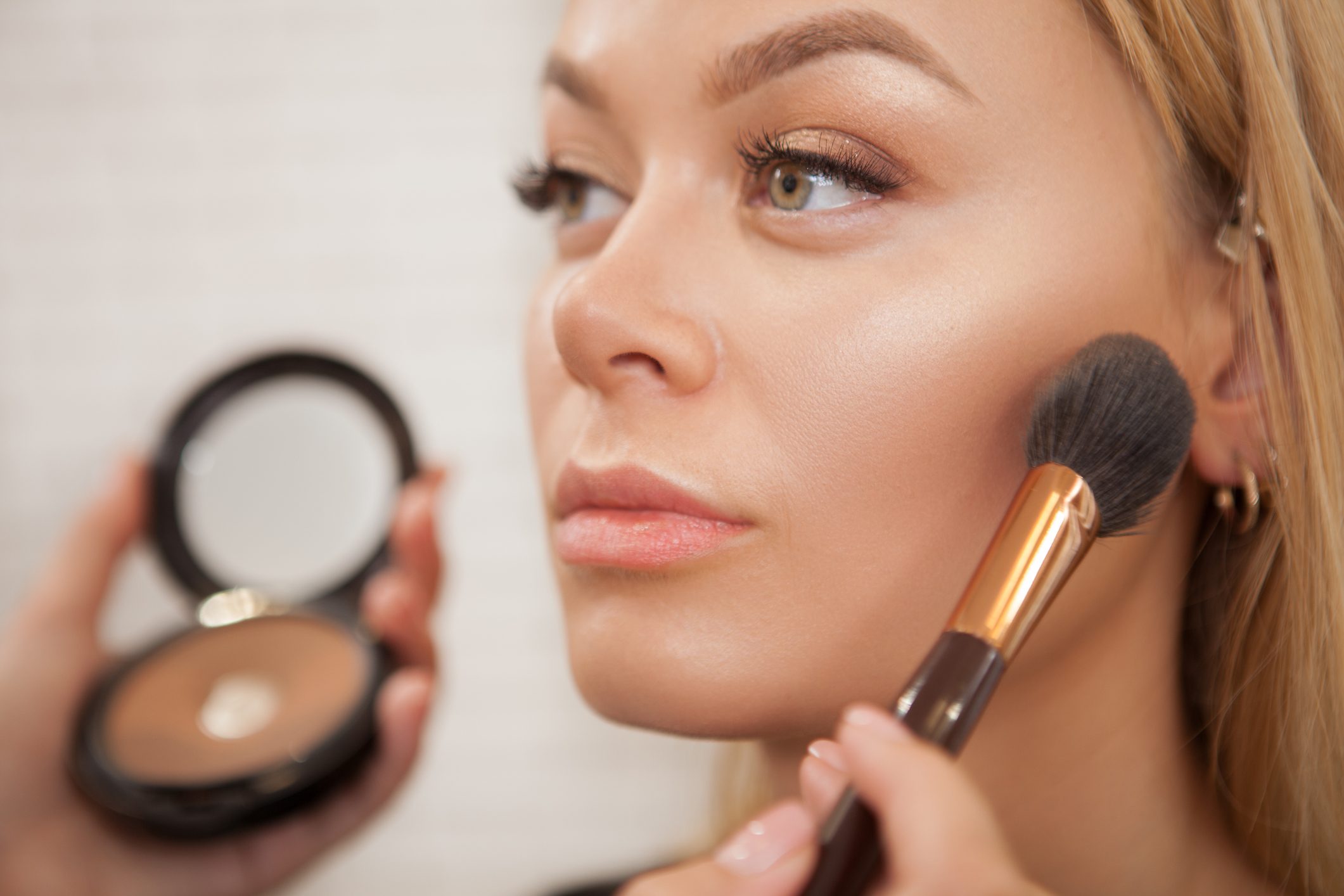 A makeup brush applies powder to a woman's cheek as she holds a compact, highlighting her face in a well-lit setting.