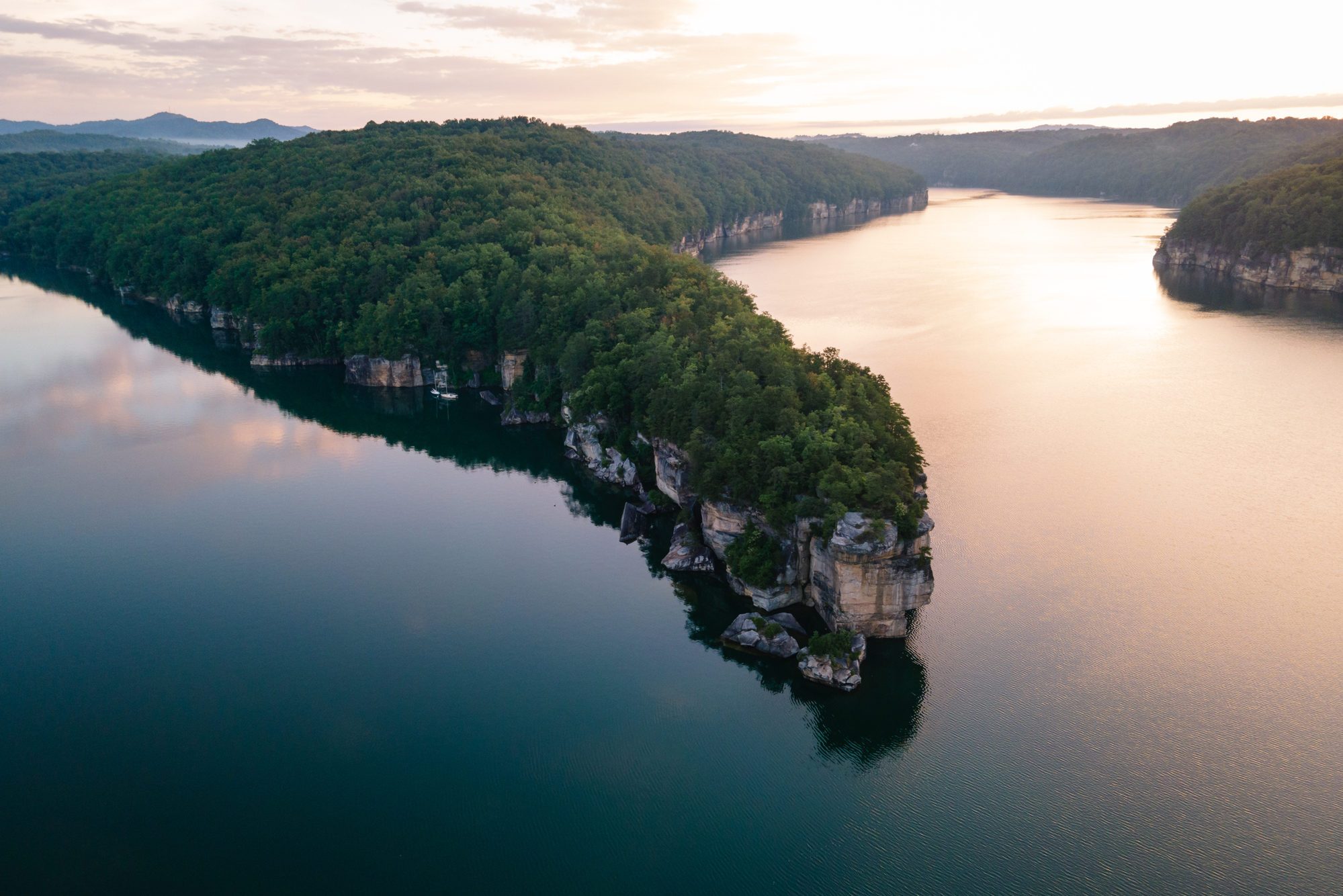Tree-covered peninsula juts into tranquil water, surrounded by rocky cliffs and distant mountains under a softly lit sky.