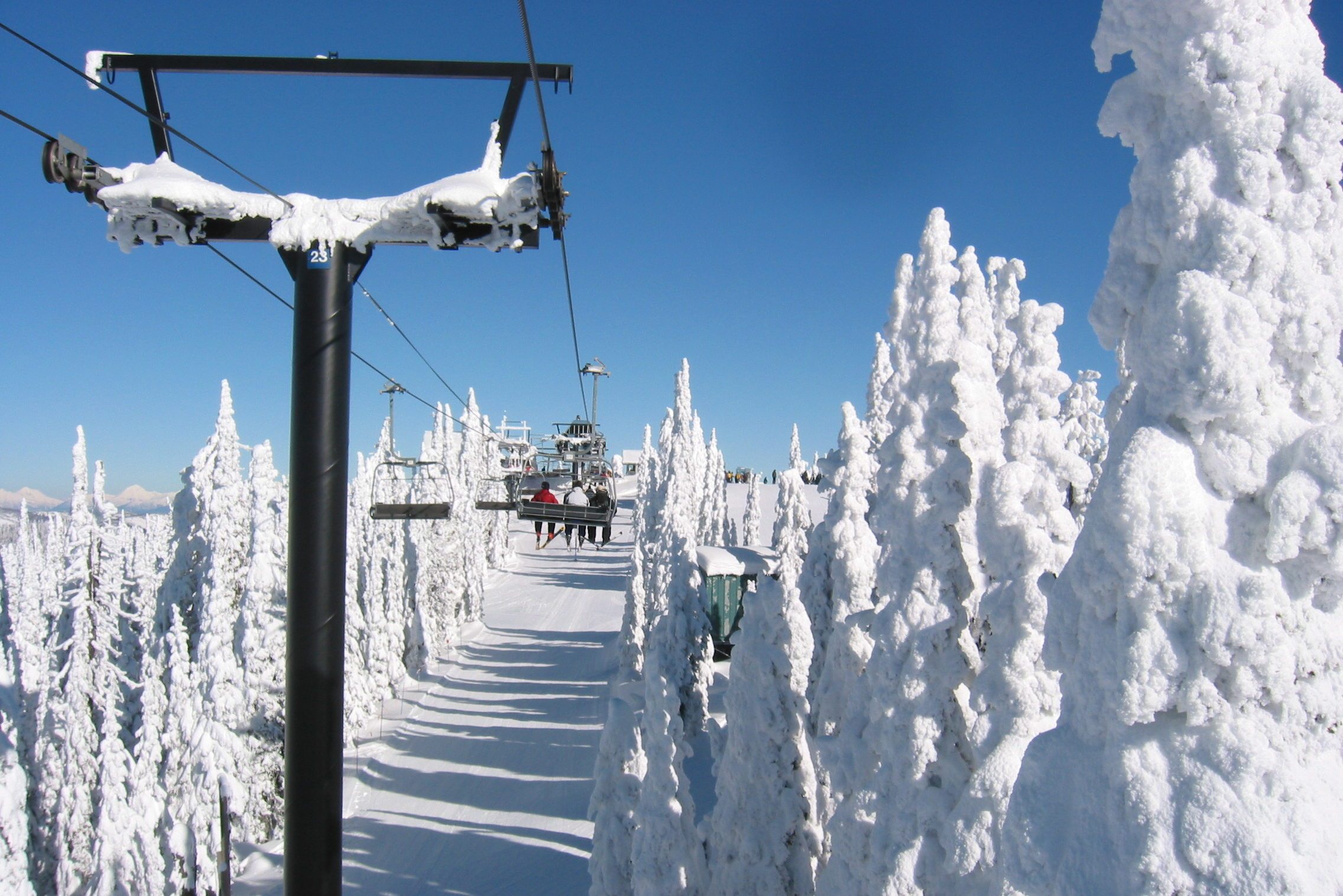 Ski lift carries people over snow-covered landscape with frosted trees under clear blue sky.