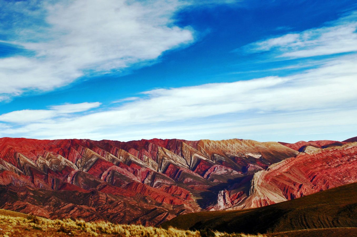 Colorful mountains form zigzag patterns, resting under a vibrant blue sky with scattered clouds. Grass-covered hills lie in the foreground.