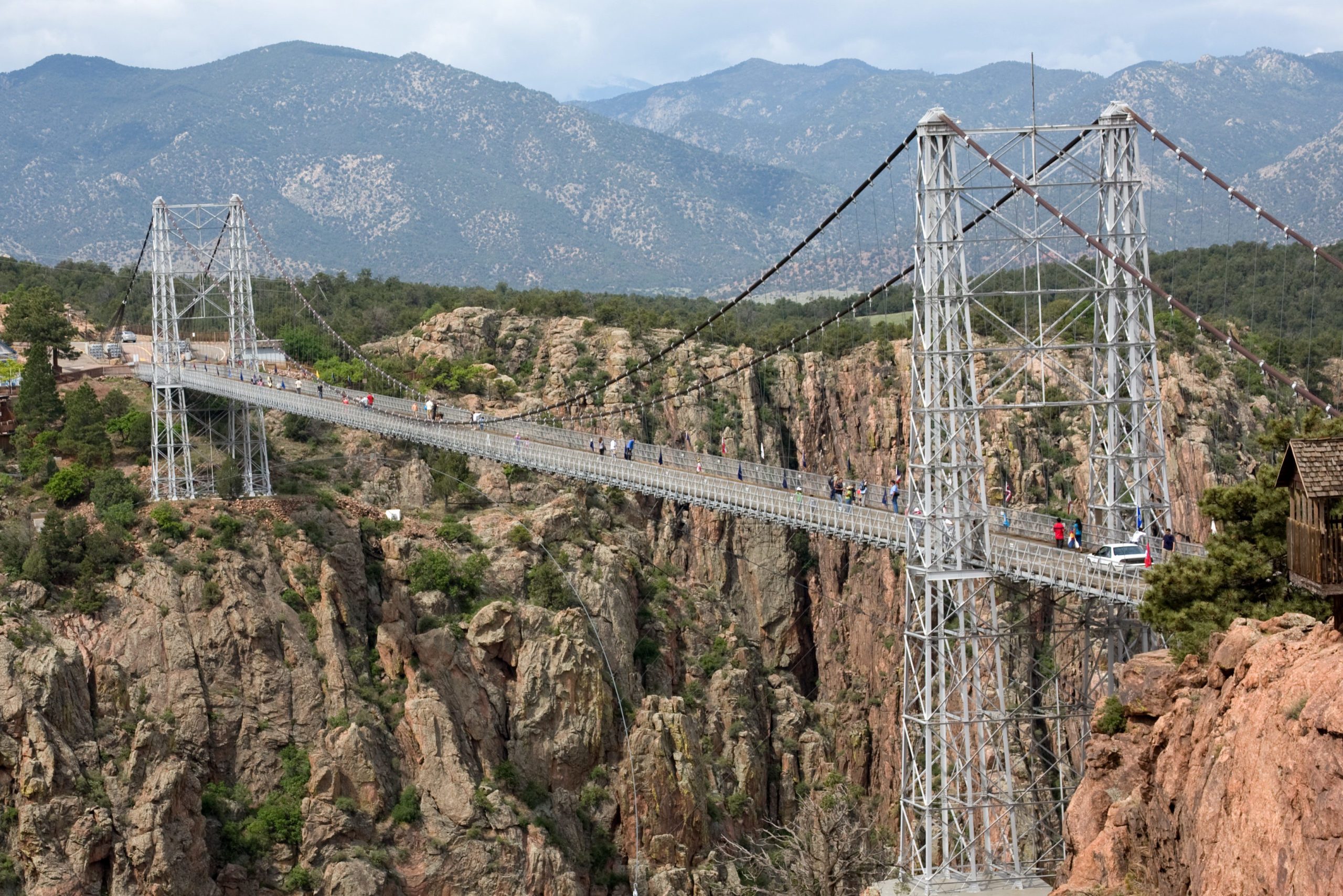 Suspension bridge spans a rocky gorge; people walk across, surrounded by lush greenery and distant mountains.