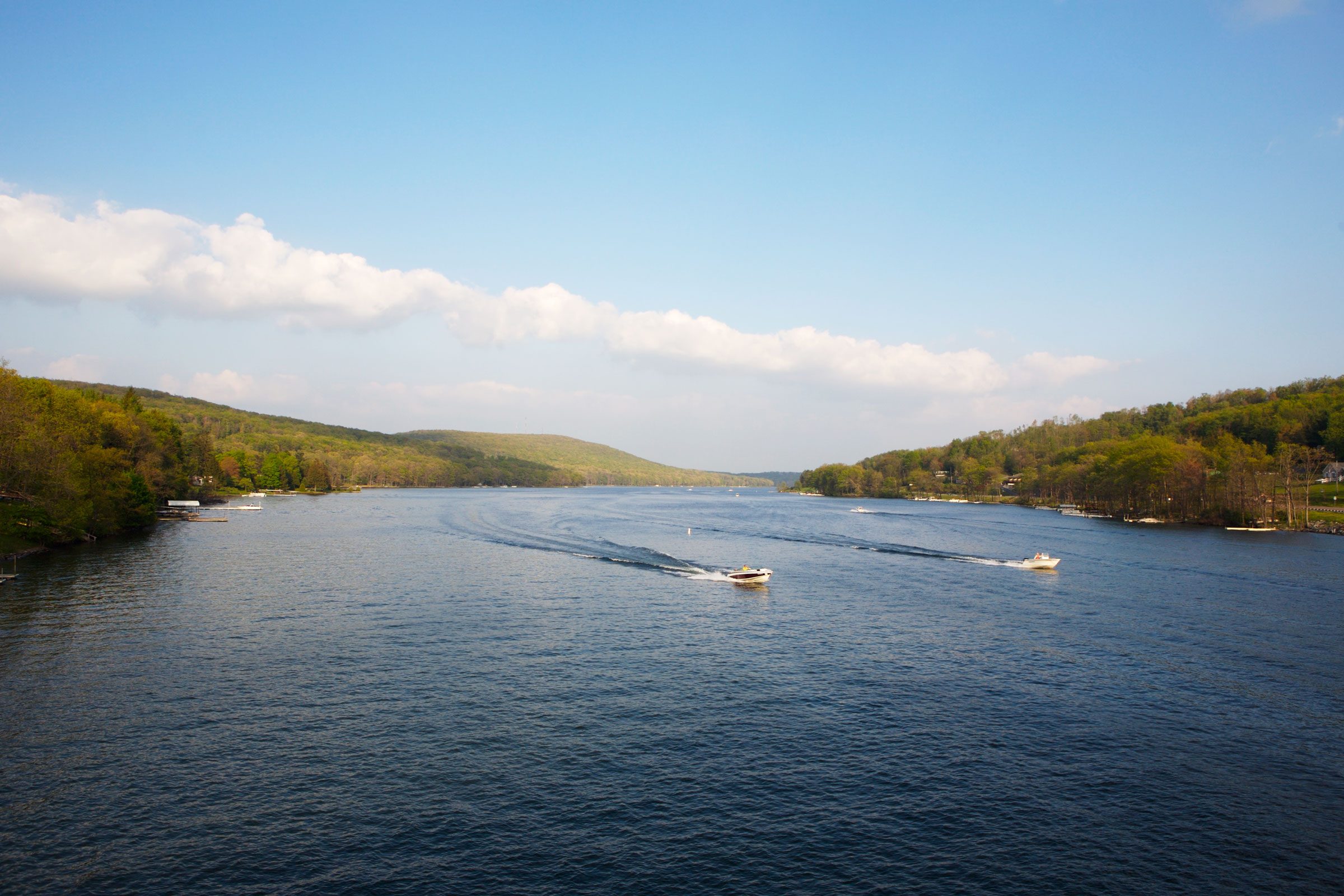 Boats create wake trails, speeding across a wide blue lake, flanked by green forested hills under a partly cloudy sky.
