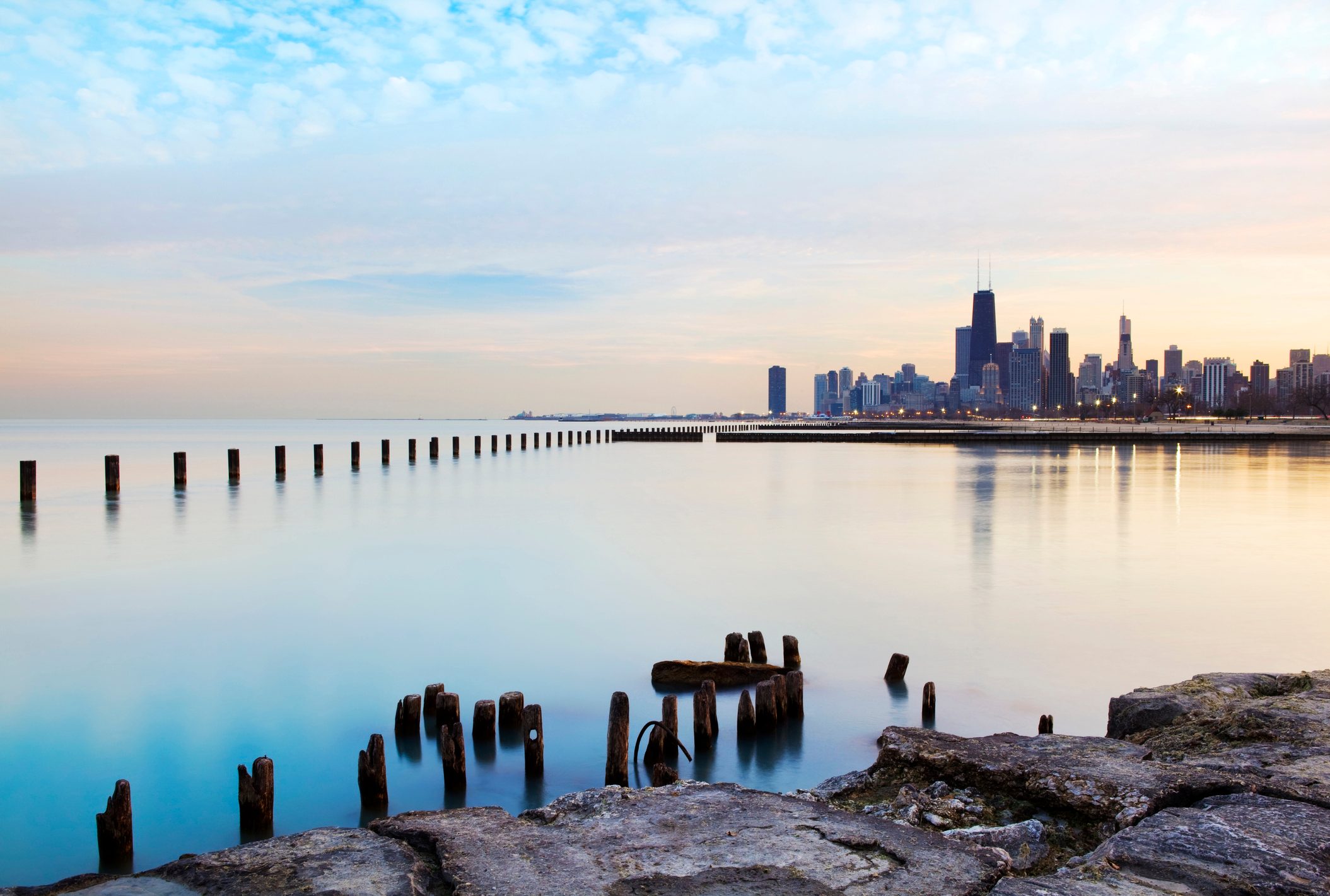 Wooden pilings stand in calm water, reflecting the city skyline under a softly clouded sky, with a rocky shore in the foreground.