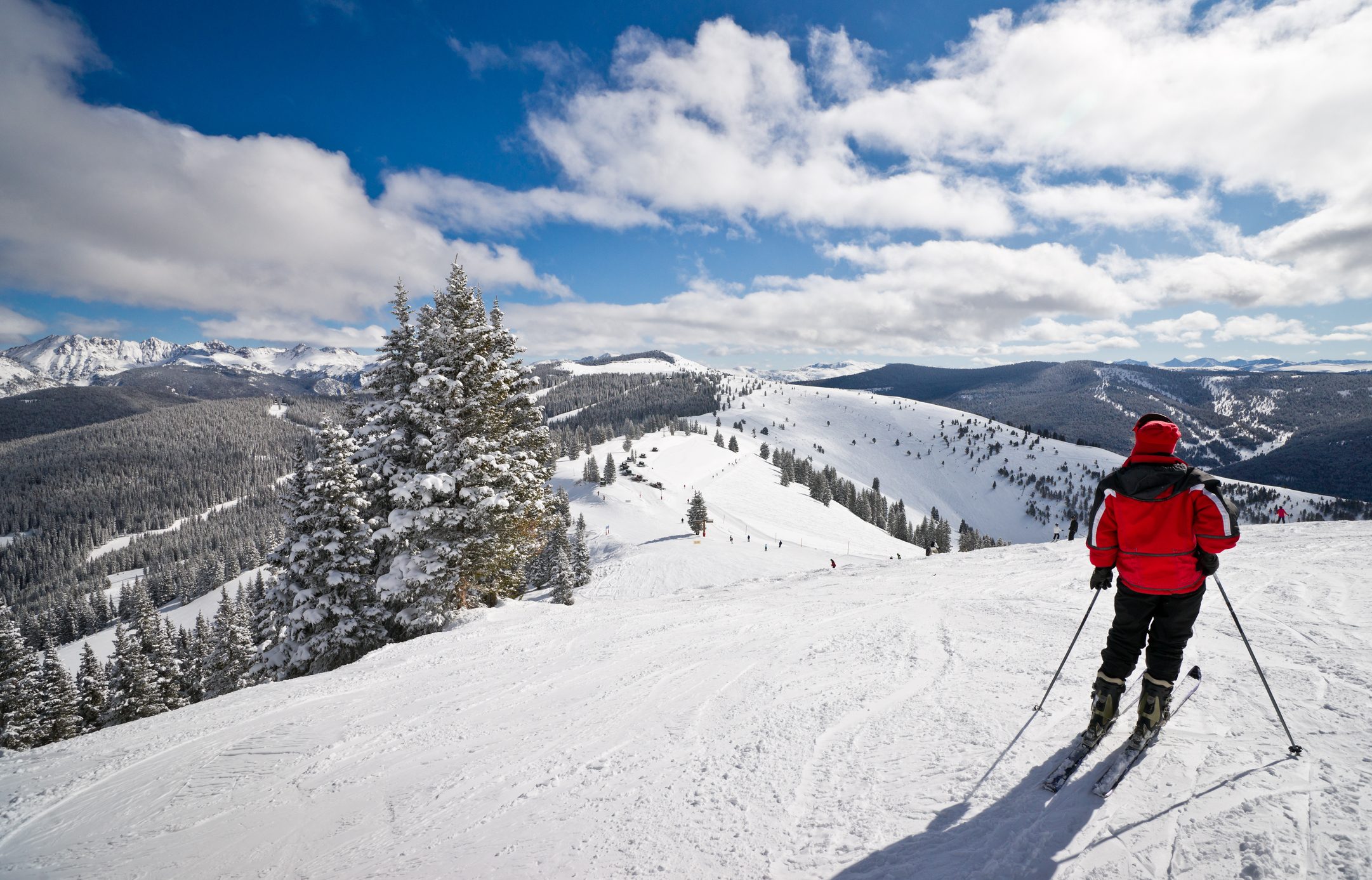 Skier stands watching on snowy slope, surrounded by picturesque mountains and pine trees under a partly cloudy blue sky.