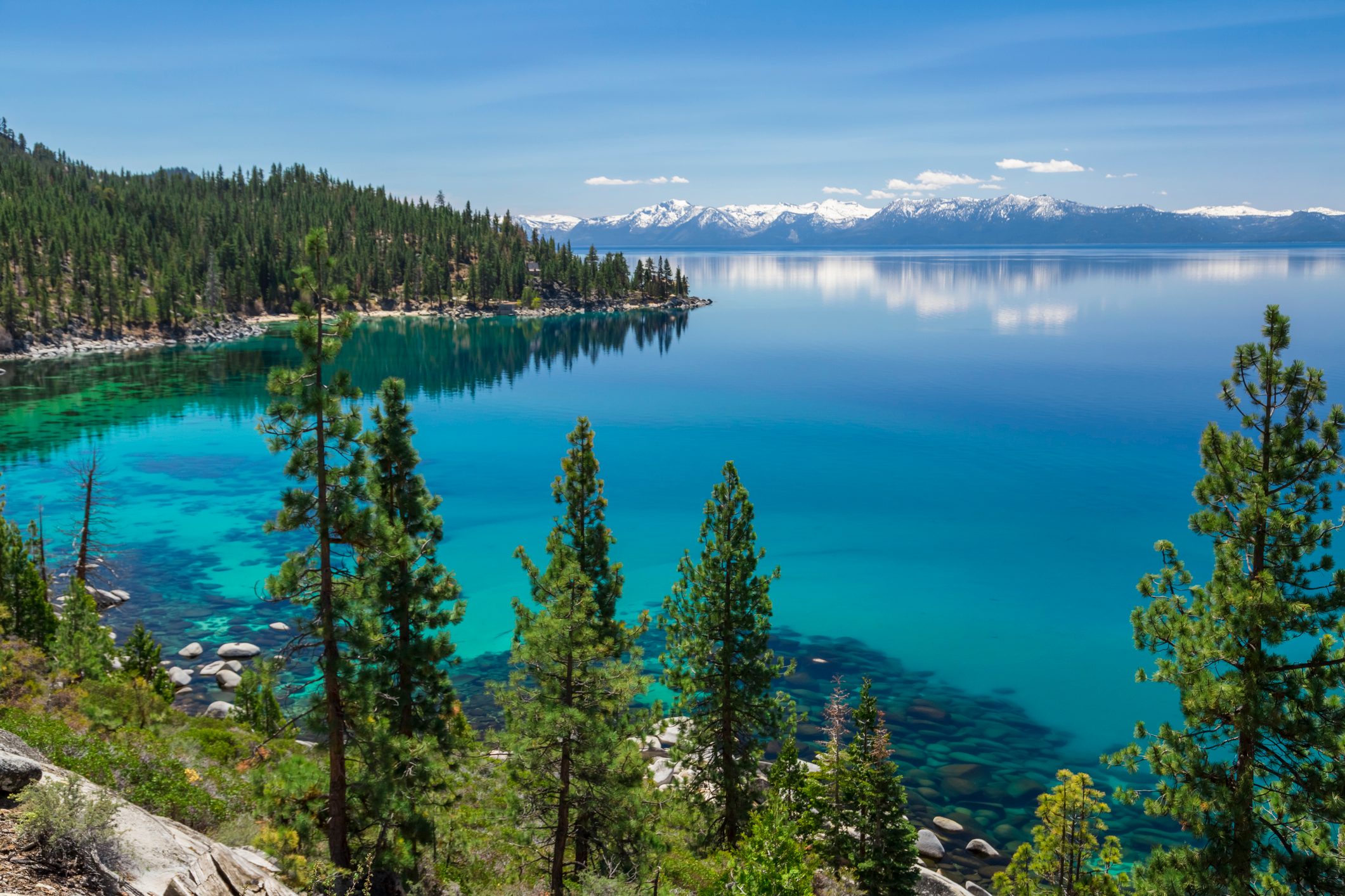 Emerald lake reflects mountains, surrounded by pine trees on rocky shore under a clear blue sky.
