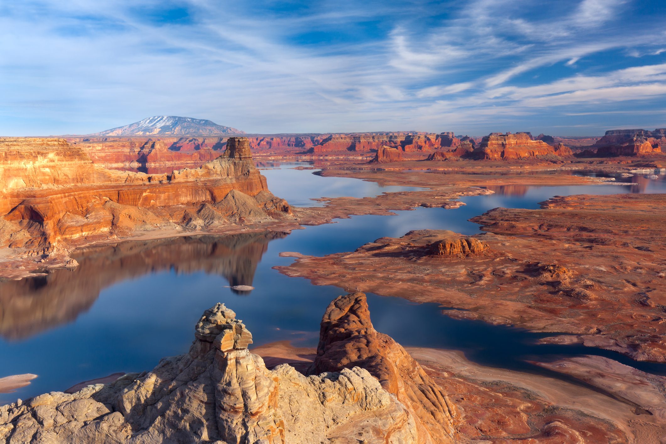 Canyon landscape with still water reflects rocky cliffs; red-orange soil and distant mountains under a blue sky create a serene, expansive scene.