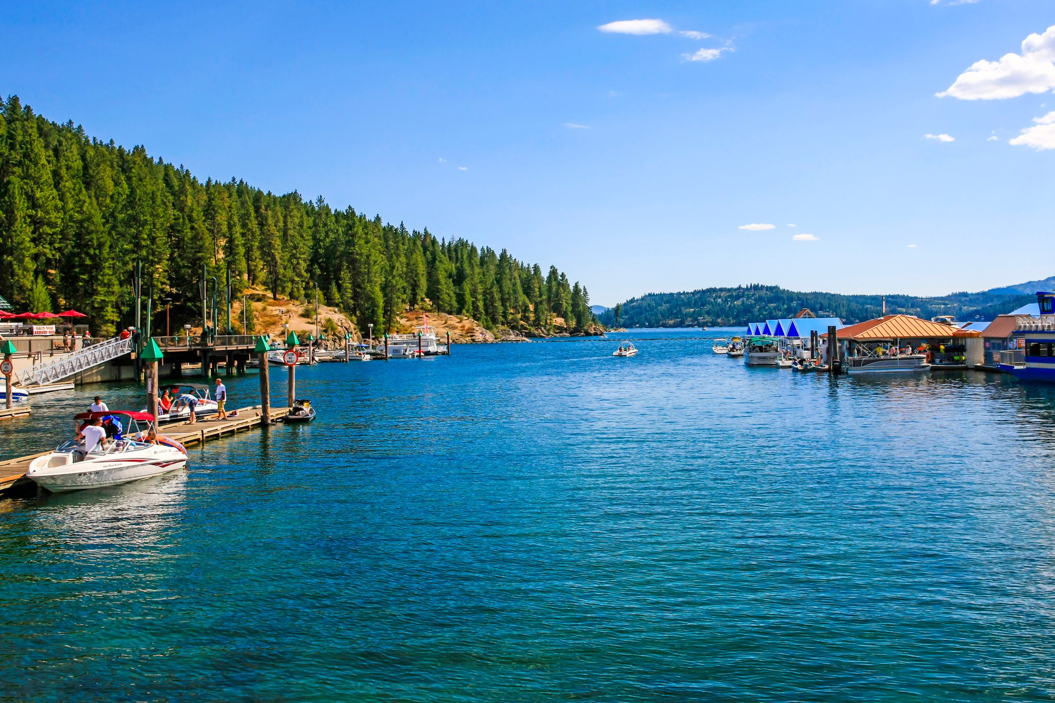 Boats dock, people socialize on a sunny marina surrounded by lush evergreen forests and calm blue water.