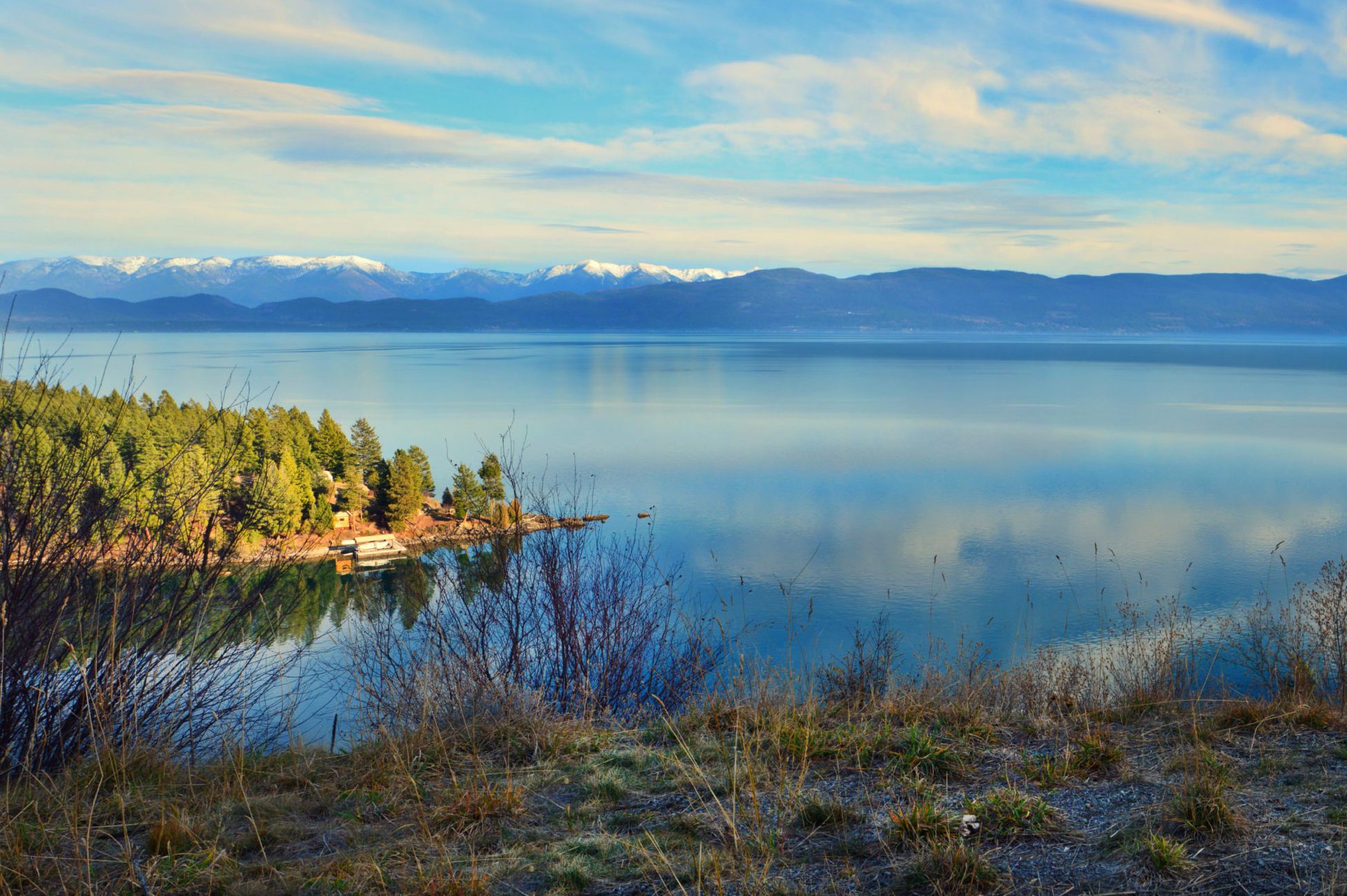 Lake reflects mountains beneath partly cloudy sky, surrounded by lush trees and grass-covered hill in serene landscape.