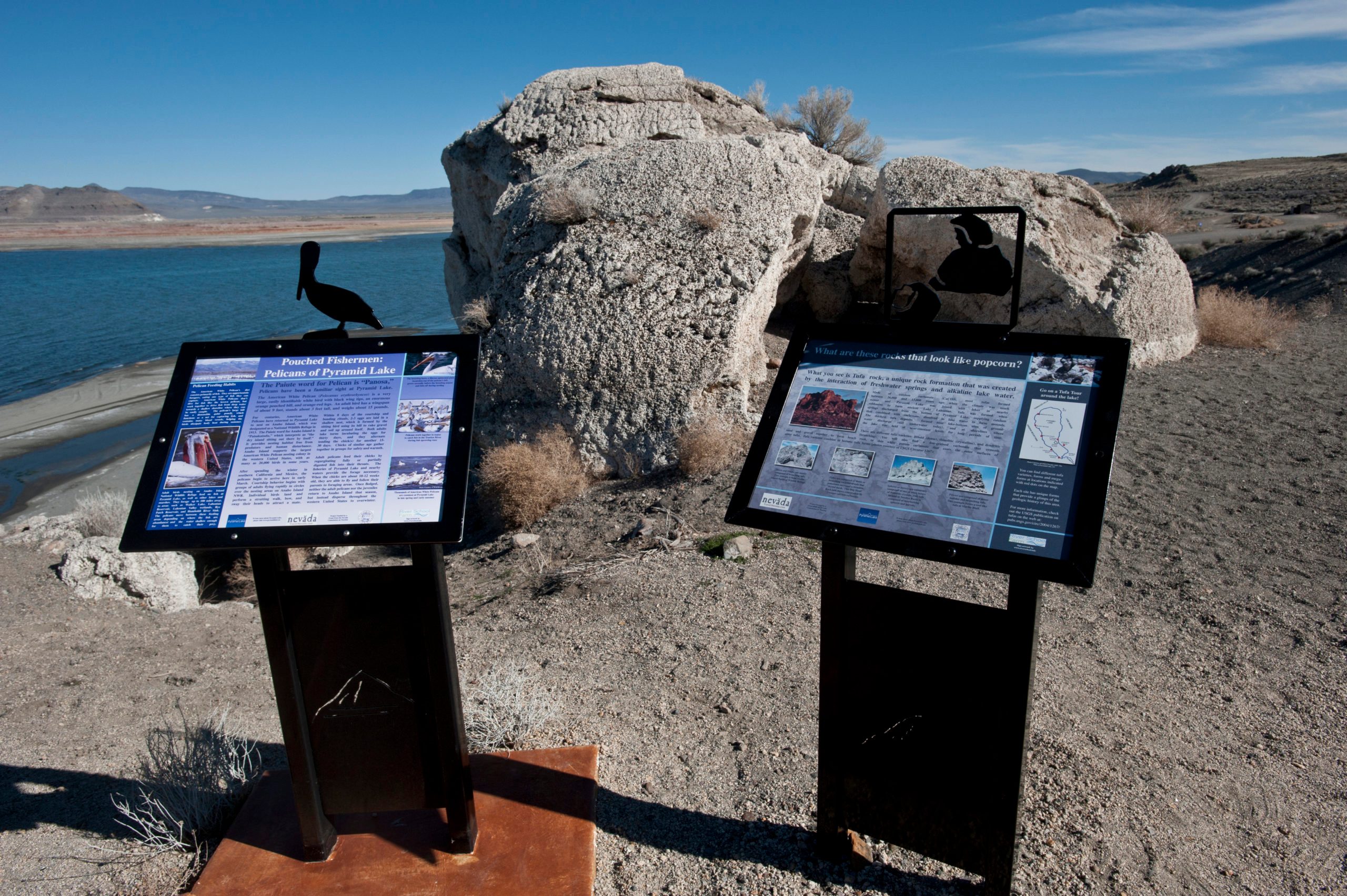 Signs display information about pelicans and rock formations. A rocky landscape with a body of water and distant mountains forms the background.