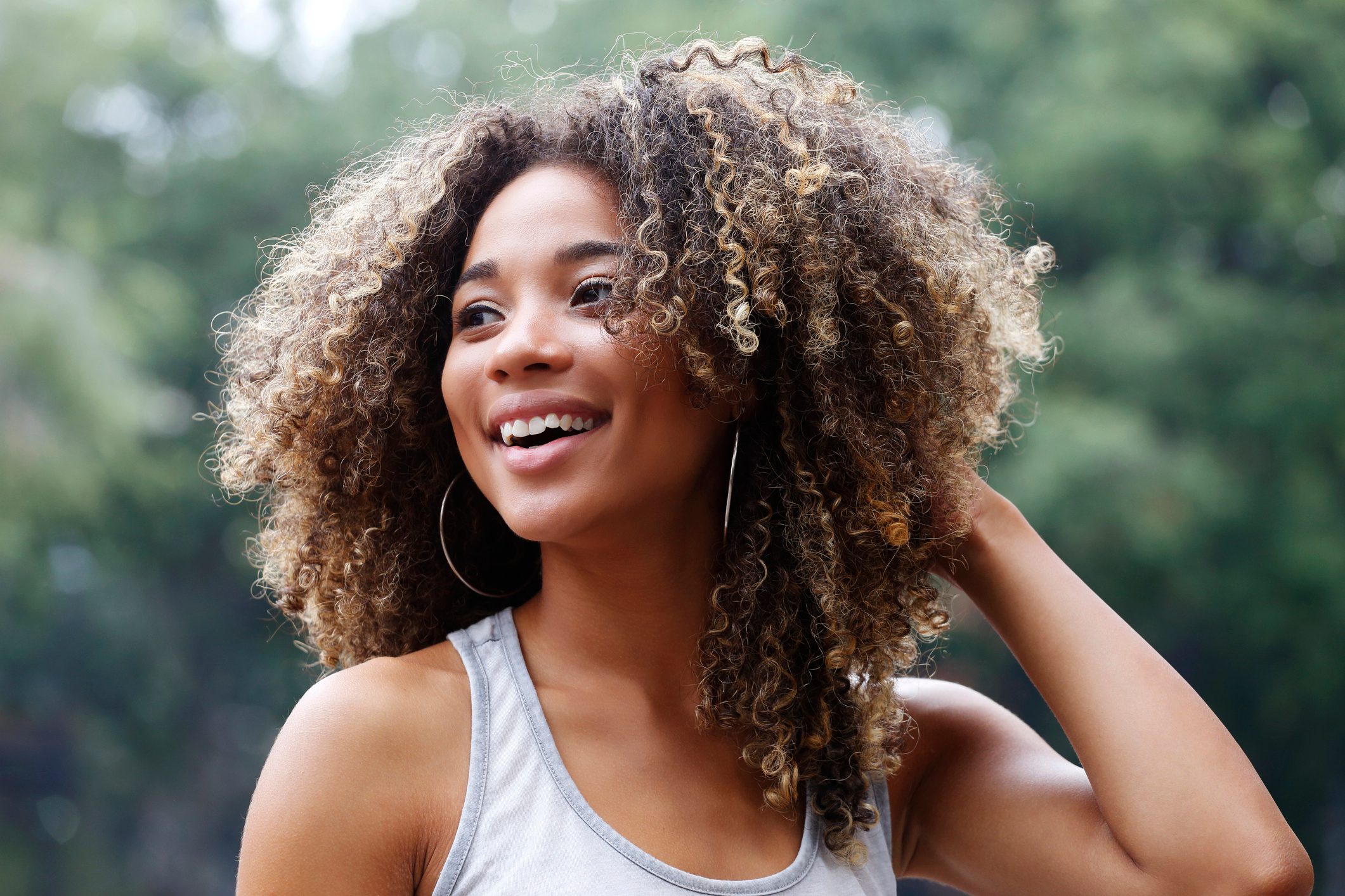 A person with curly hair smiles while touching their head, standing in a green, outdoor setting.