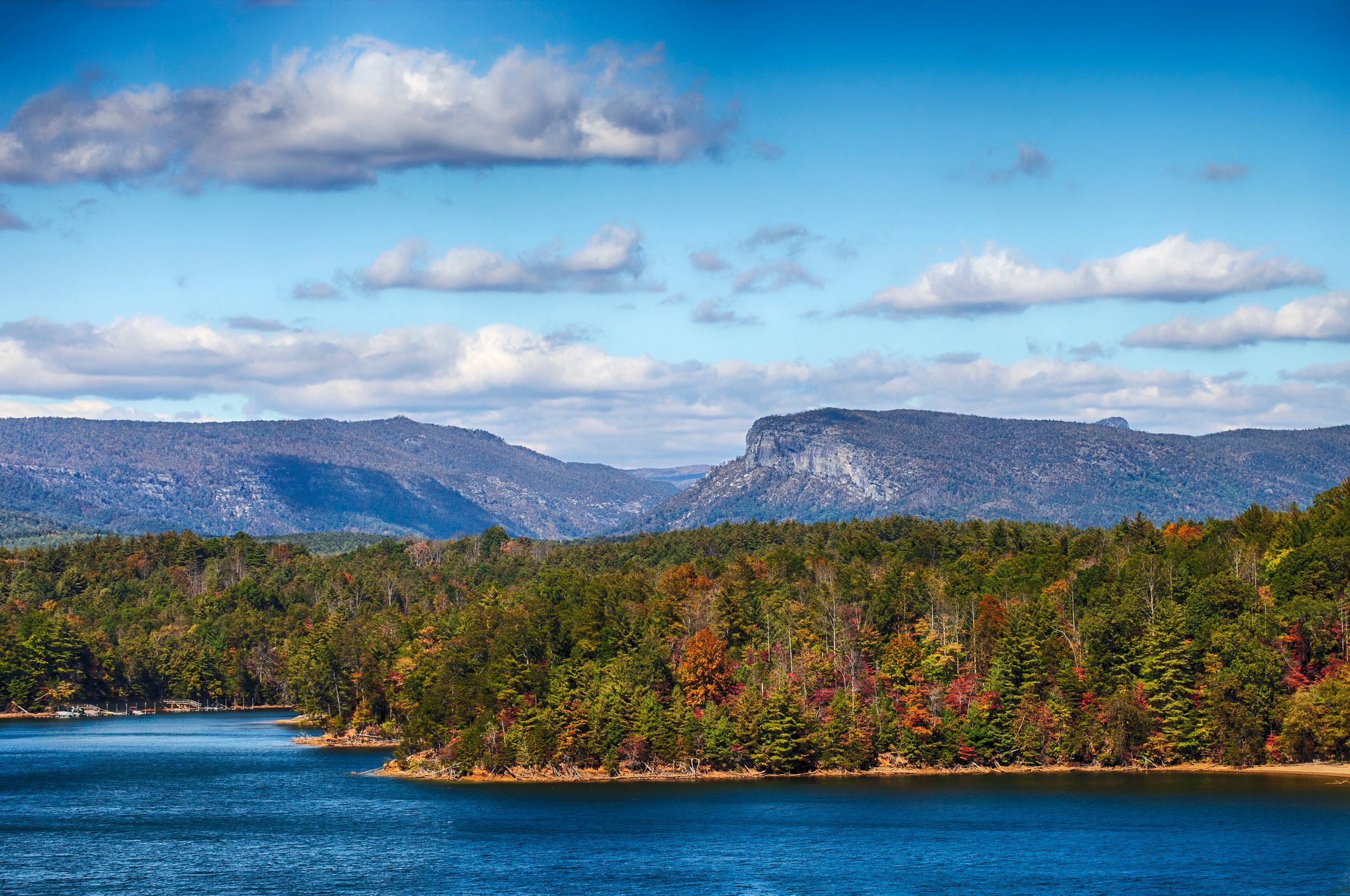 Mountains overlook a forested shoreline bordering a blue lake under a partly cloudy sky.