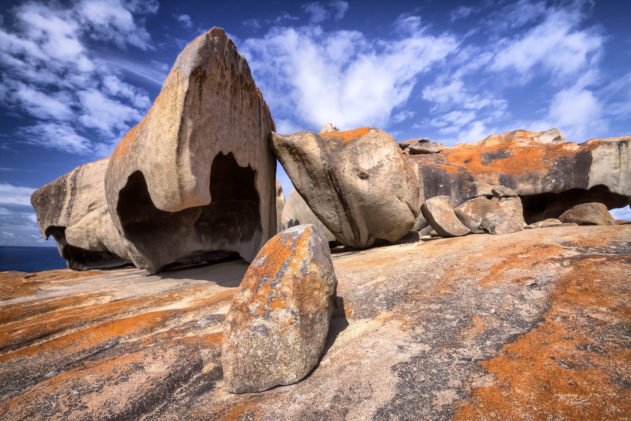 Large, weathered rocks rest on a sunlit, flat terrain under a partly cloudy blue sky, showcasing unique shapes and orange lichen patches.