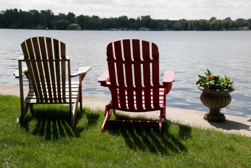 Two Adirondack chairs face a calm lake. The chairs are on grassy terrain, with a stone planter holding flowers nearby.