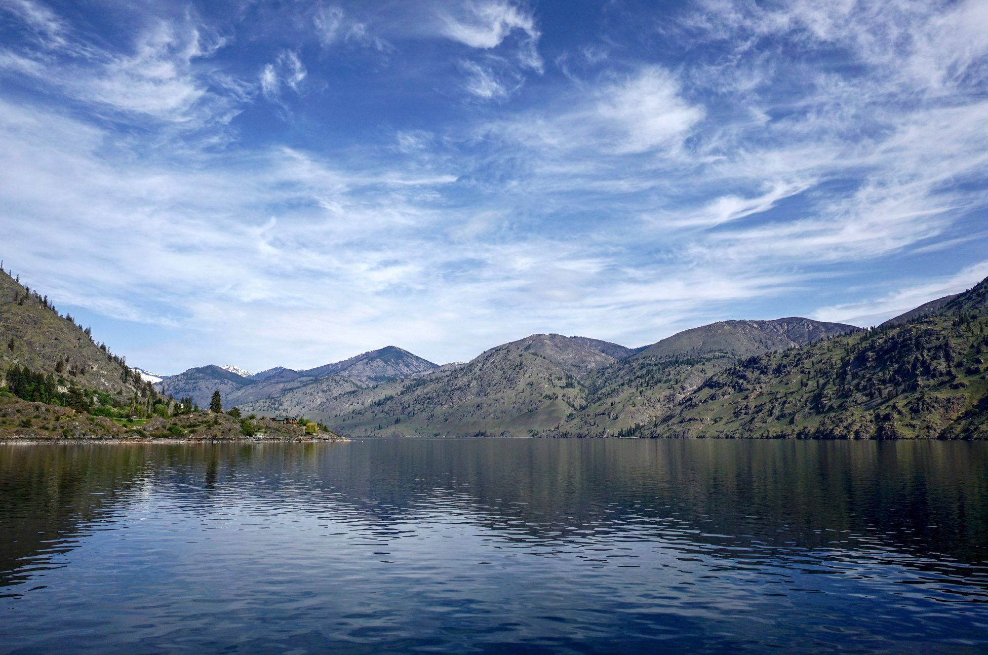 Calm lake reflects mountainous landscape, surrounded by greenery under a partly cloudy sky.