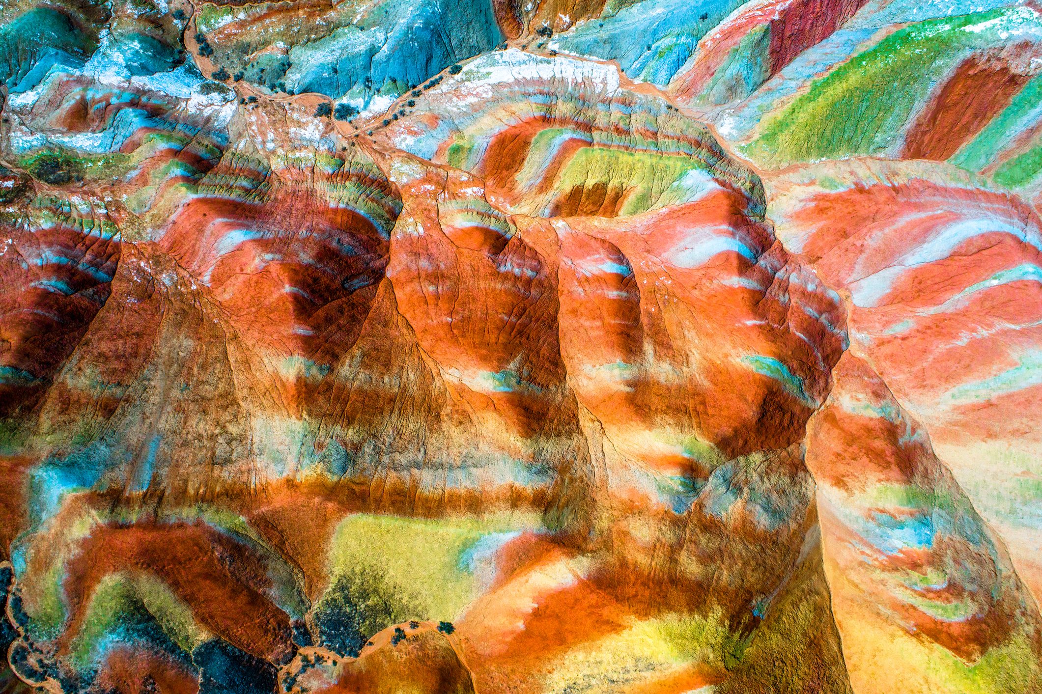 Colorful rock formations display vibrant layers; red, orange, blue, green, and yellow hues blend seamlessly, creating a surreal landscape from an aerial perspective.