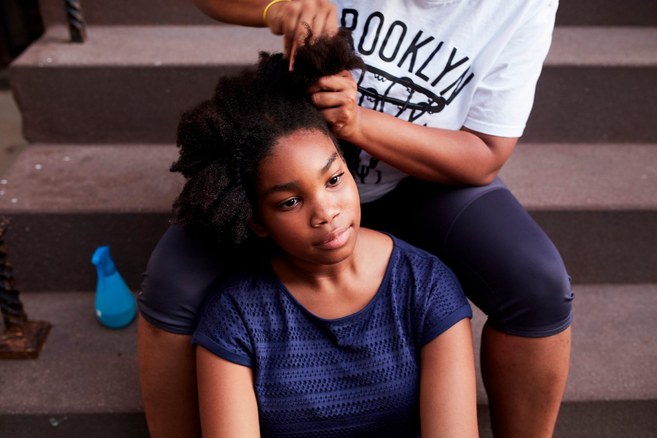 Girl sitting on stairs, having hair styled by another person; blue bottle nearby, casual atmosphere.