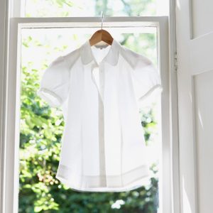 White shirt hangs on a wooden hanger inside a bright window, with green foliage visible outside.