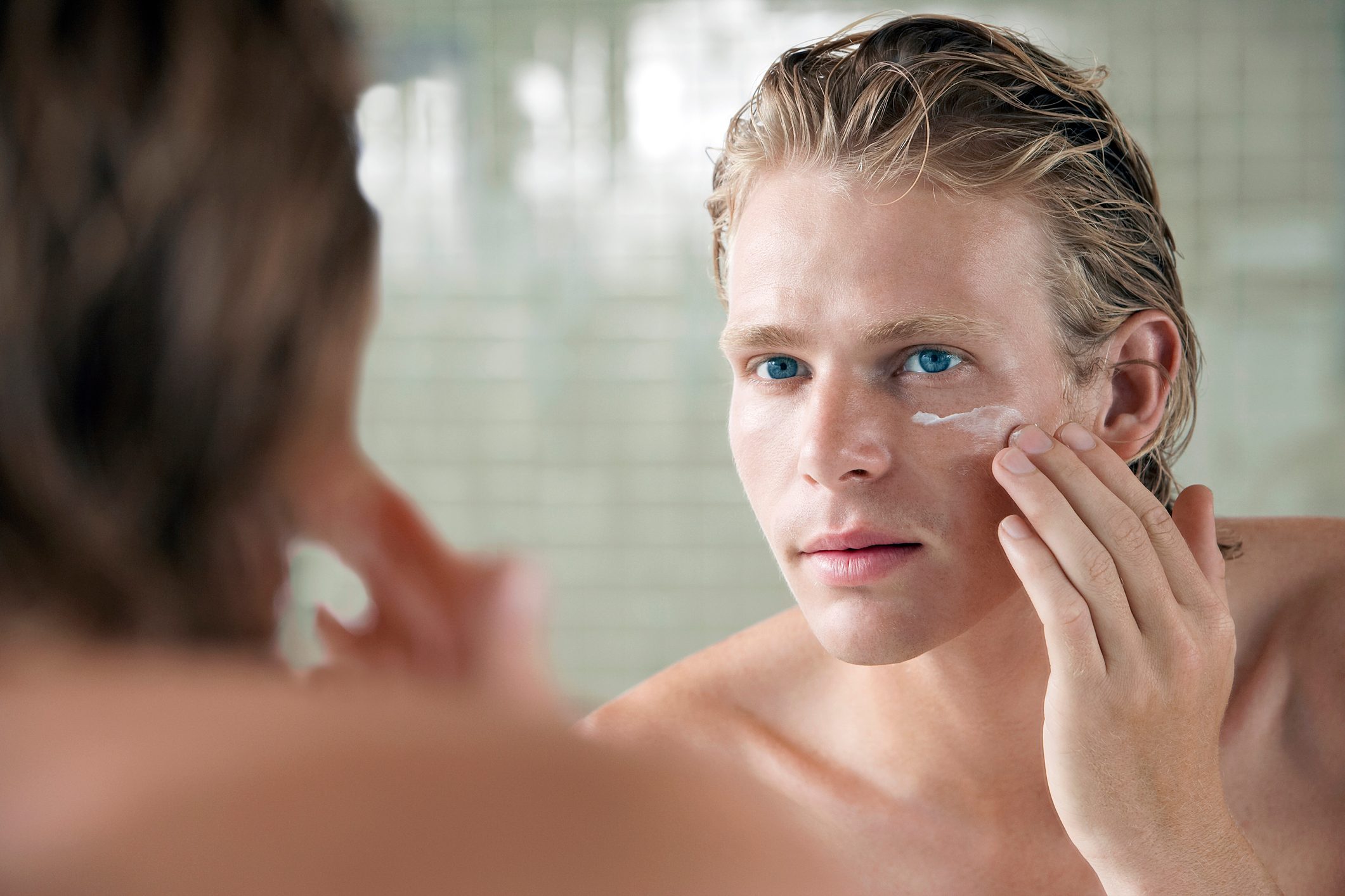 A person applies cream to their face while looking at a bathroom mirror, tiles visible in the background.