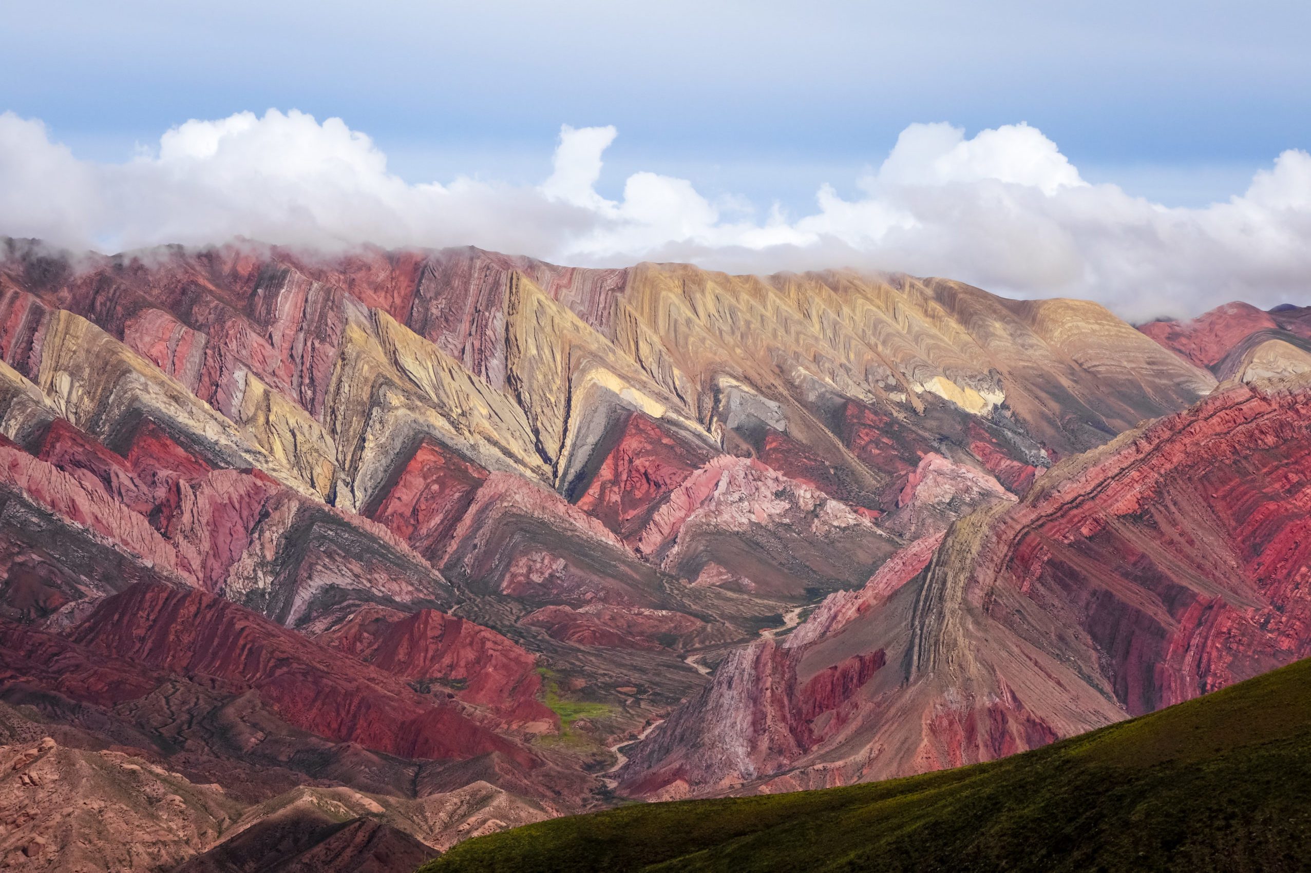 Colorful mountains display vibrant, layered stripes with red, pink, yellow hues beneath cloudy sky, creating a striking visual effect in a rugged landscape.
