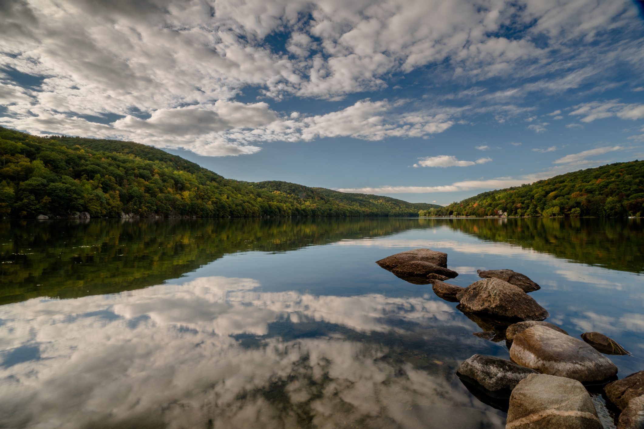 Rocks form a path in a calm lake, reflecting the cloudy sky, surrounded by forested hills.