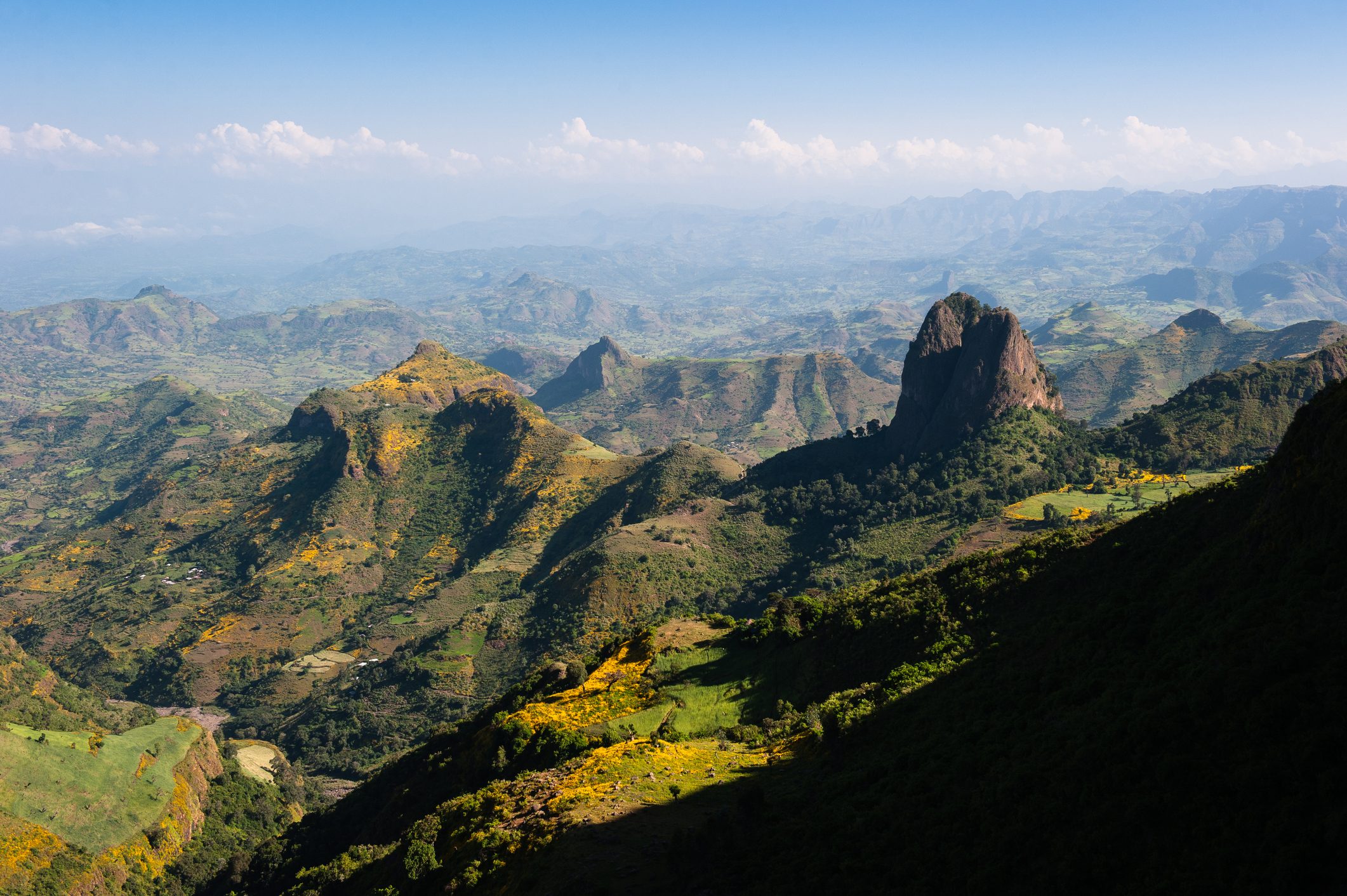 Mountains with greenery sit under clear blue skies, casting shadows across the valley below, creating a dramatic landscape.