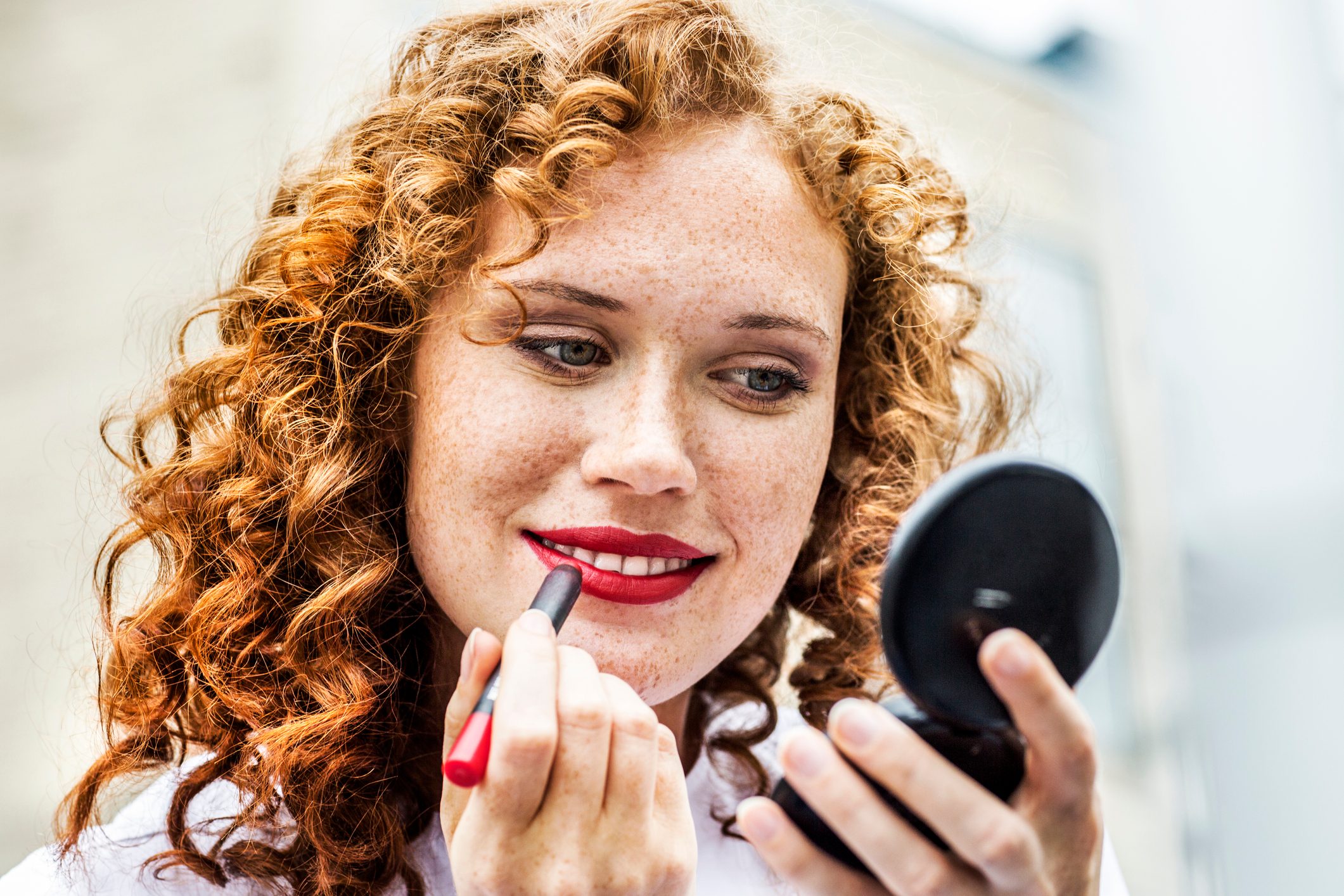 A person applies red lipstick while holding a compact mirror, outdoors with a blurred background.