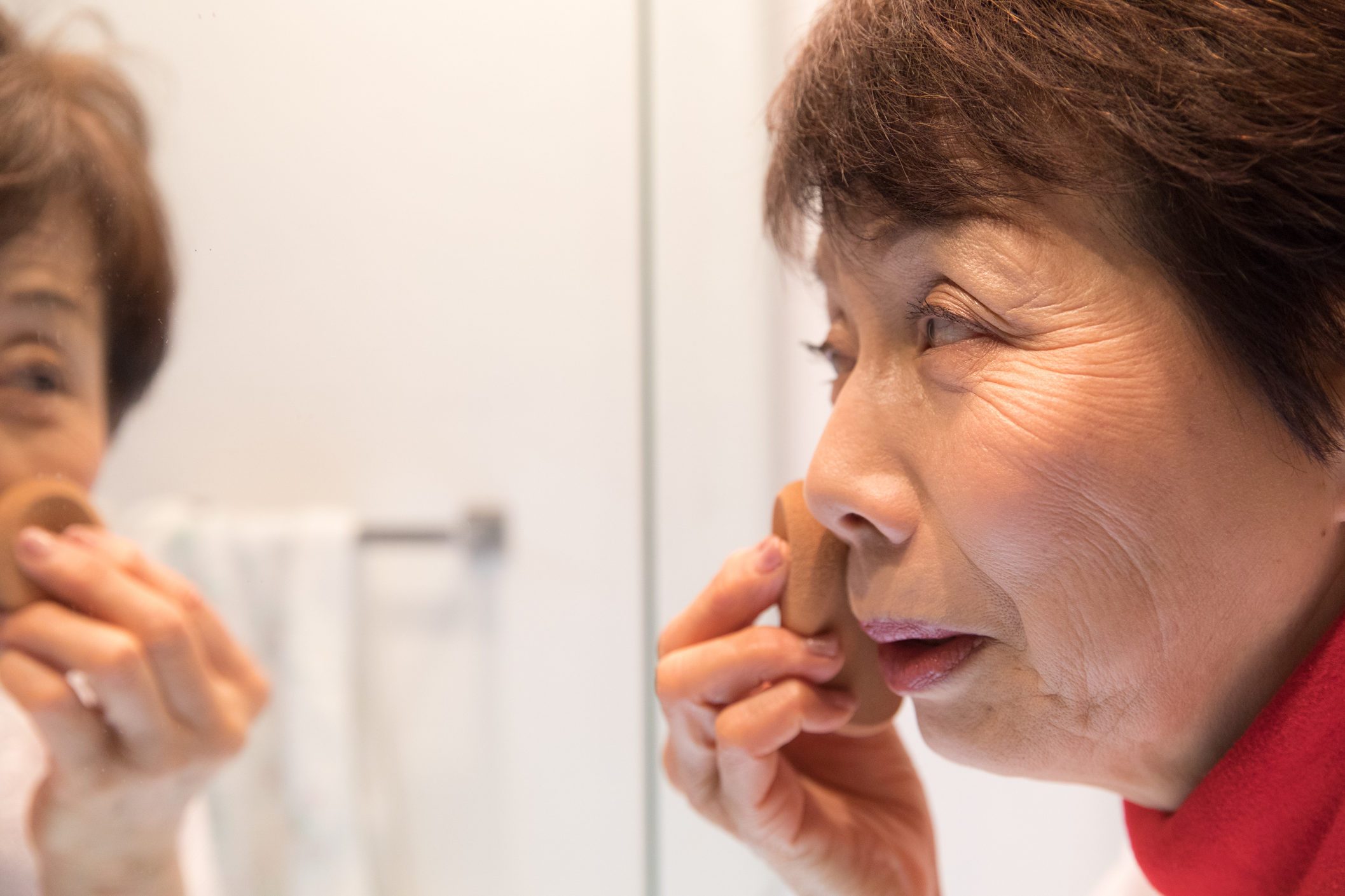 Person applying makeup sponge, gazing into a bathroom mirror, with blurred white towel visible in the background.