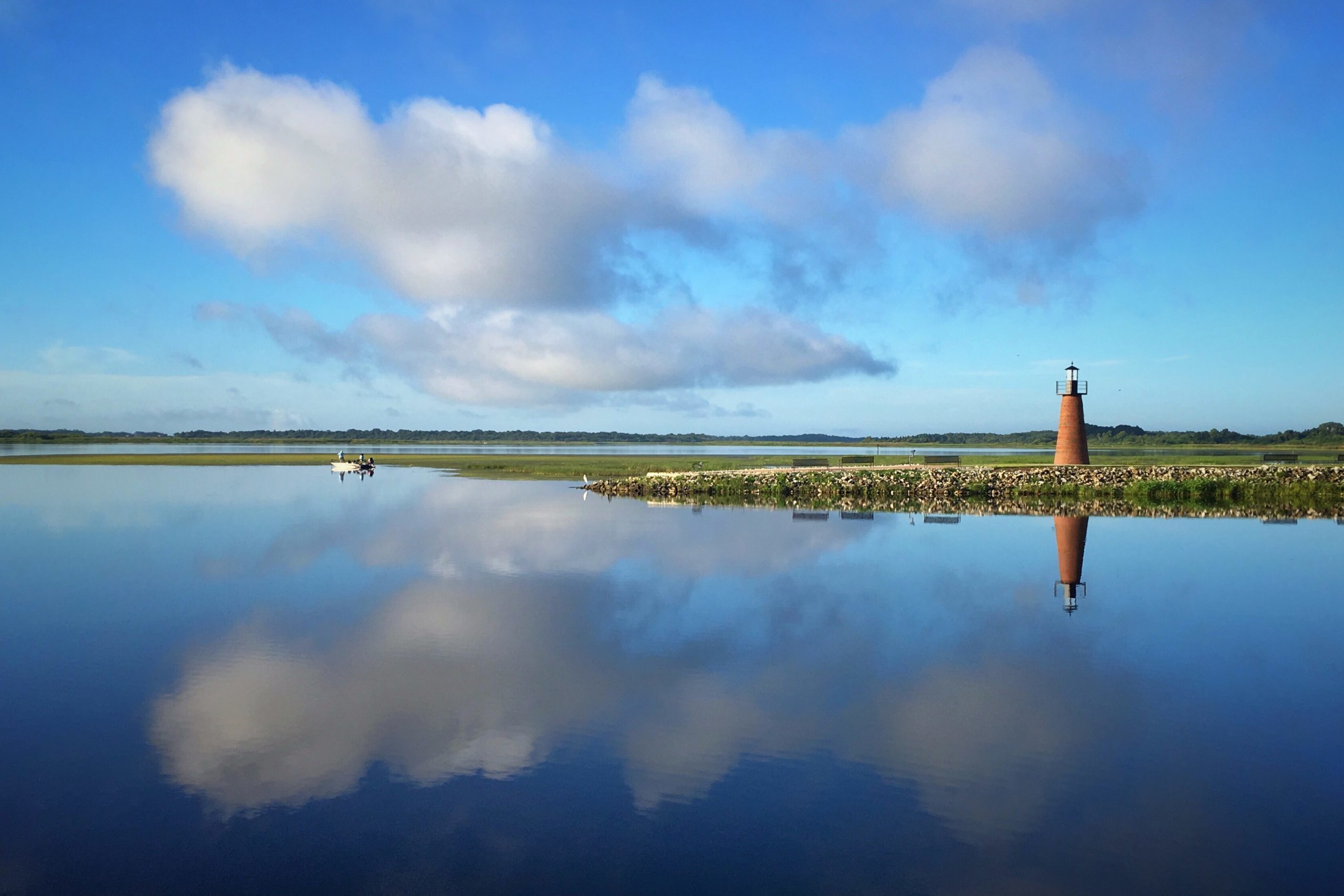 A lighthouse reflects in calm water, surrounded by a grassy landscape under a bright blue sky with scattered clouds.