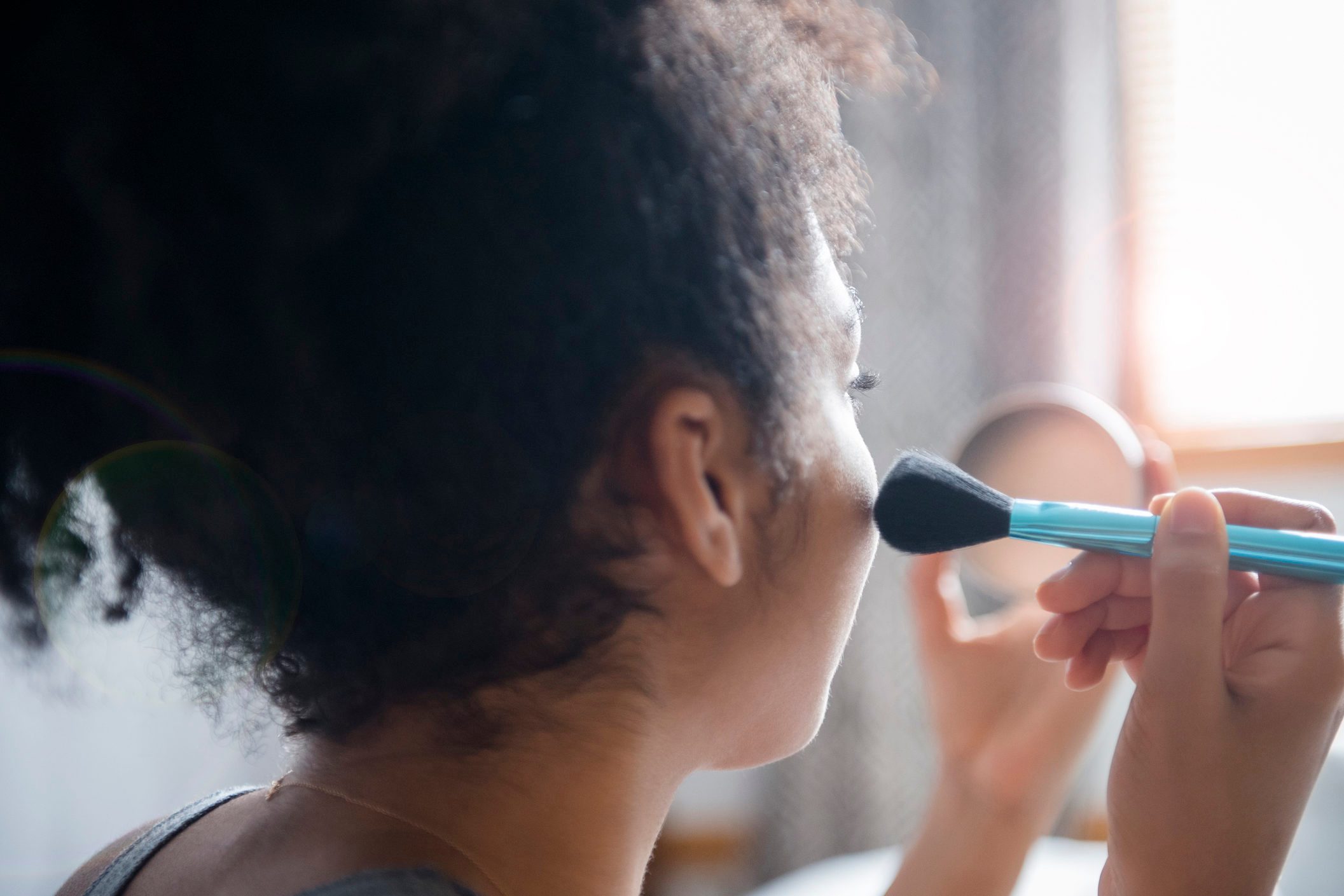 Person applies makeup with a brush, holding a mirror, in a softly lit room.
