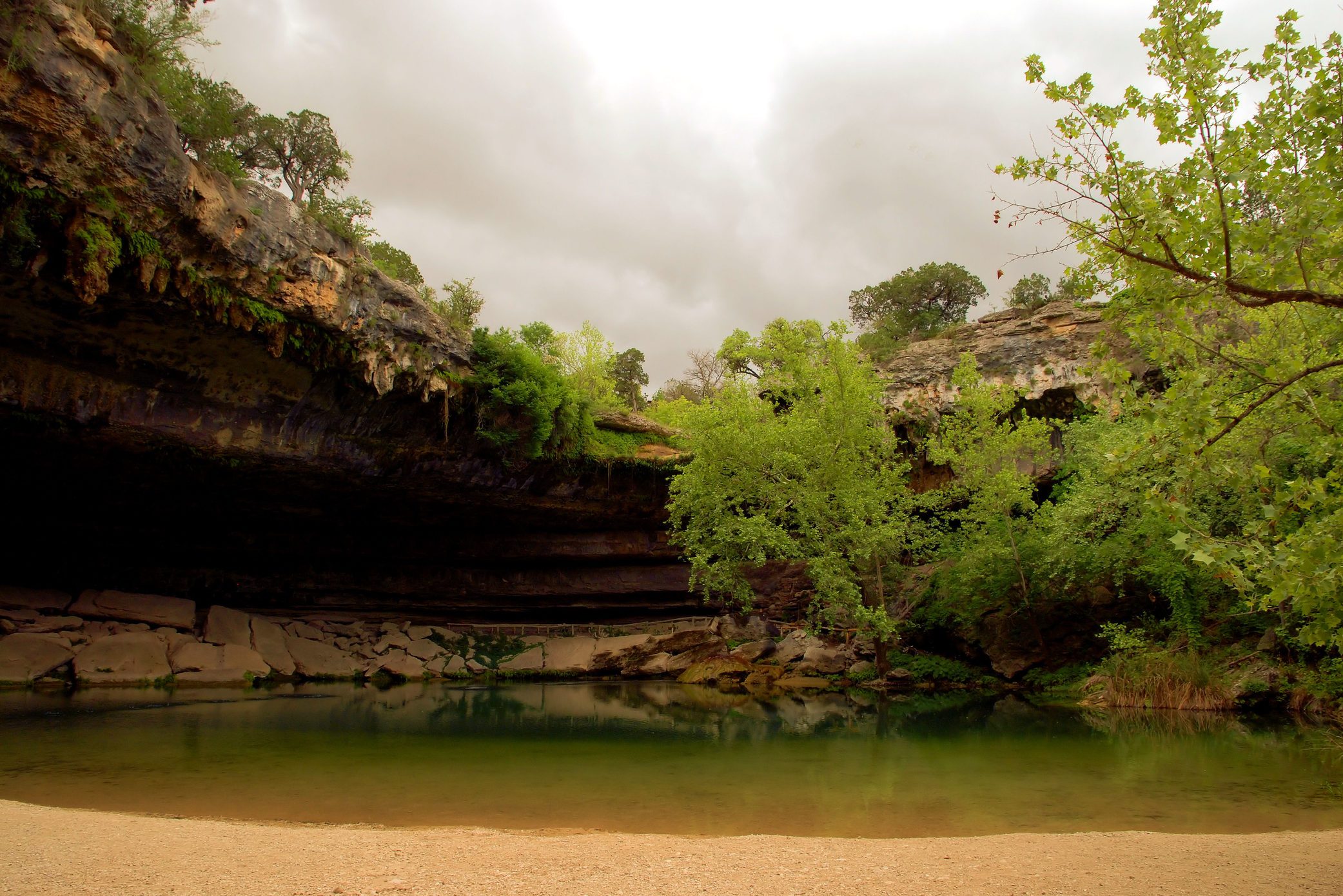 A serene pool reflects rocky overhangs and lush greenery under an overcast sky, creating a tranquil, natural oasis.