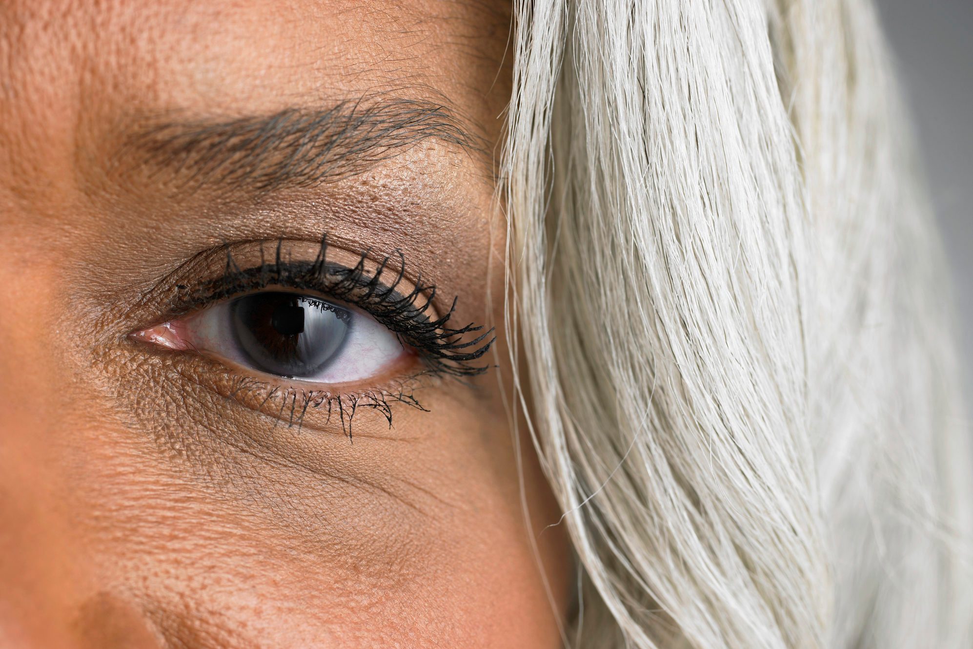 An eye with mascara adorns a face framed by gray hair, detailed against a plain background.