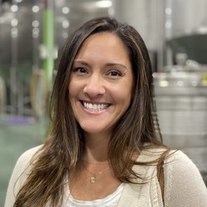 Woman smiling indoors, standing in an industrial setting with large metal tanks and green accents in the background.