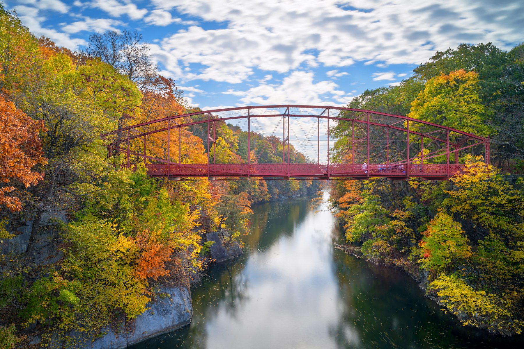 Red bridge spans a calm river, surrounded by trees with vibrant autumn leaves, under a partly cloudy blue sky.