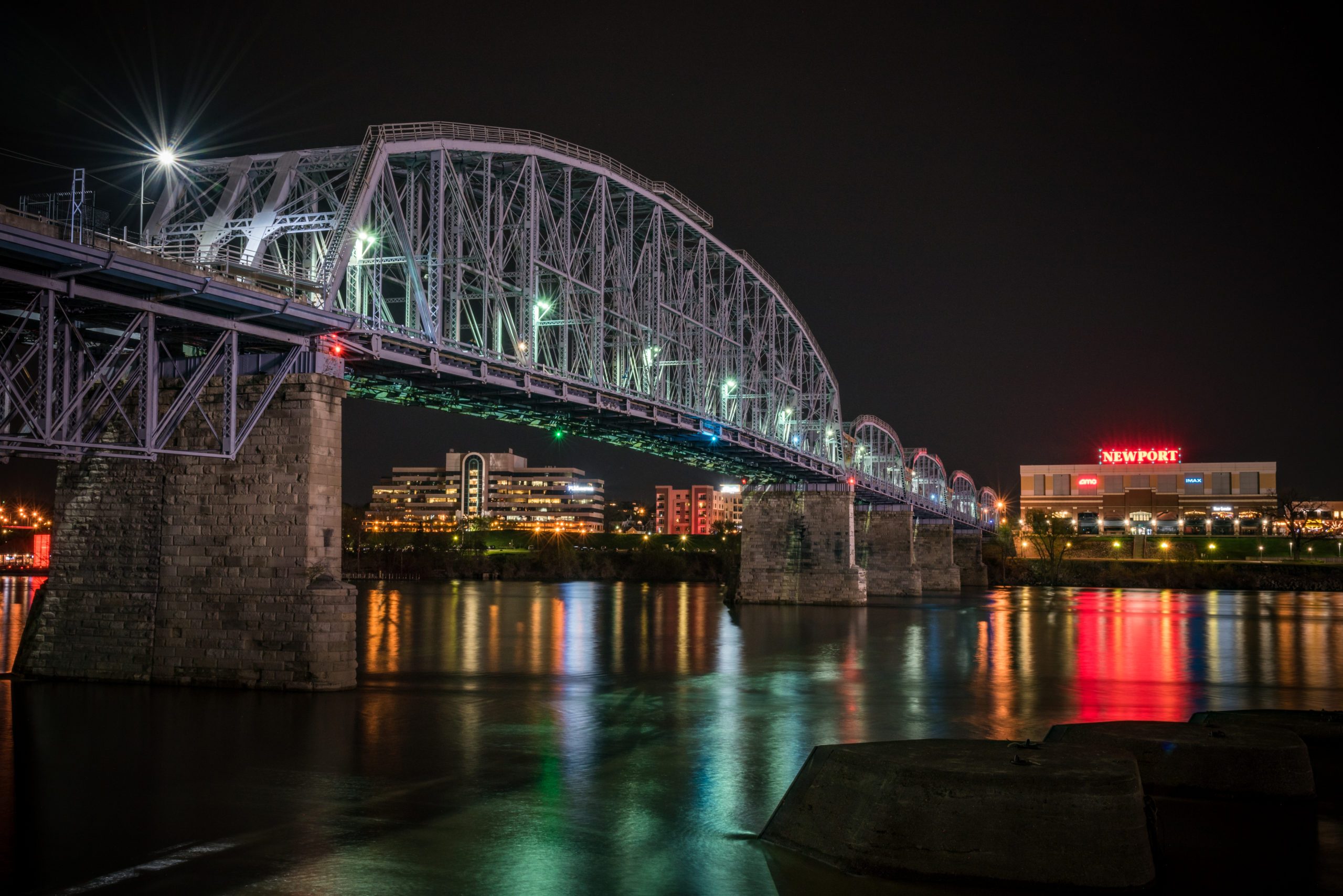 Bridge spans river, illuminated at night; city buildings with a red "Newport" sign in the background reflect colorful lights in the water.