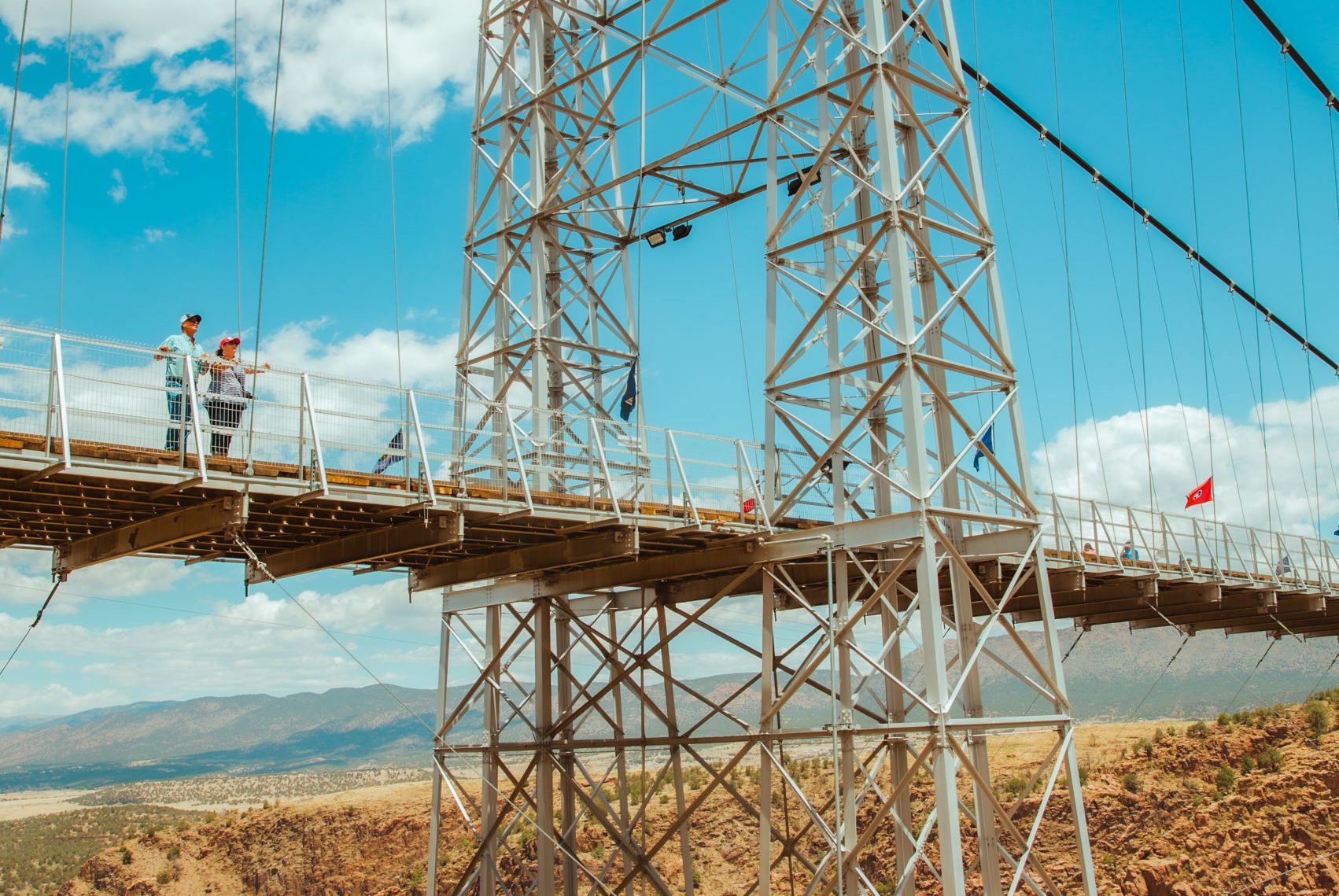 People stand on a suspension bridge, overlooking a canyon under a blue sky with scattered clouds. A red flag is visible on the bridge.